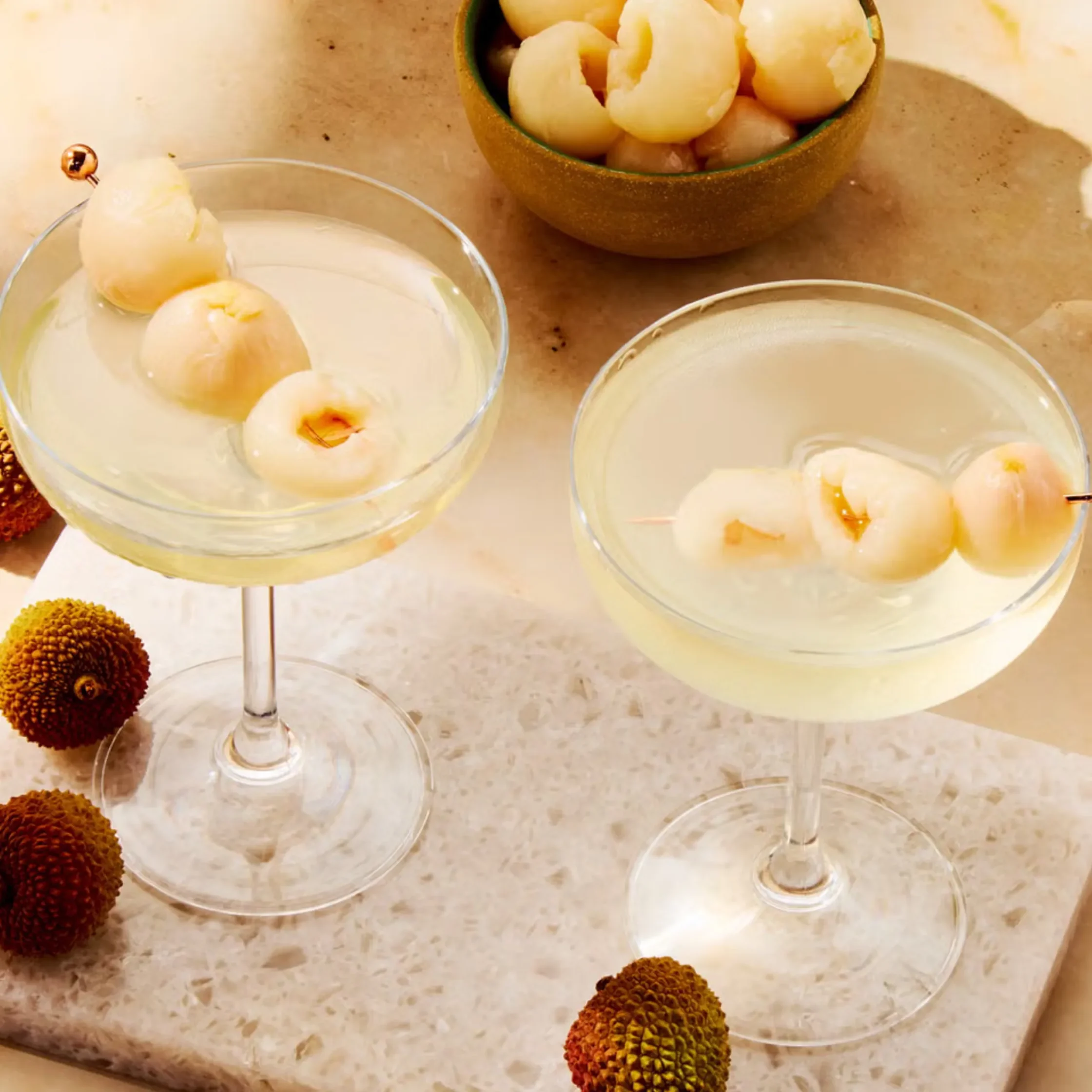 Cocktail glasses with pale lychee garnishes and a bowl of peeled lychee on a marble surface.