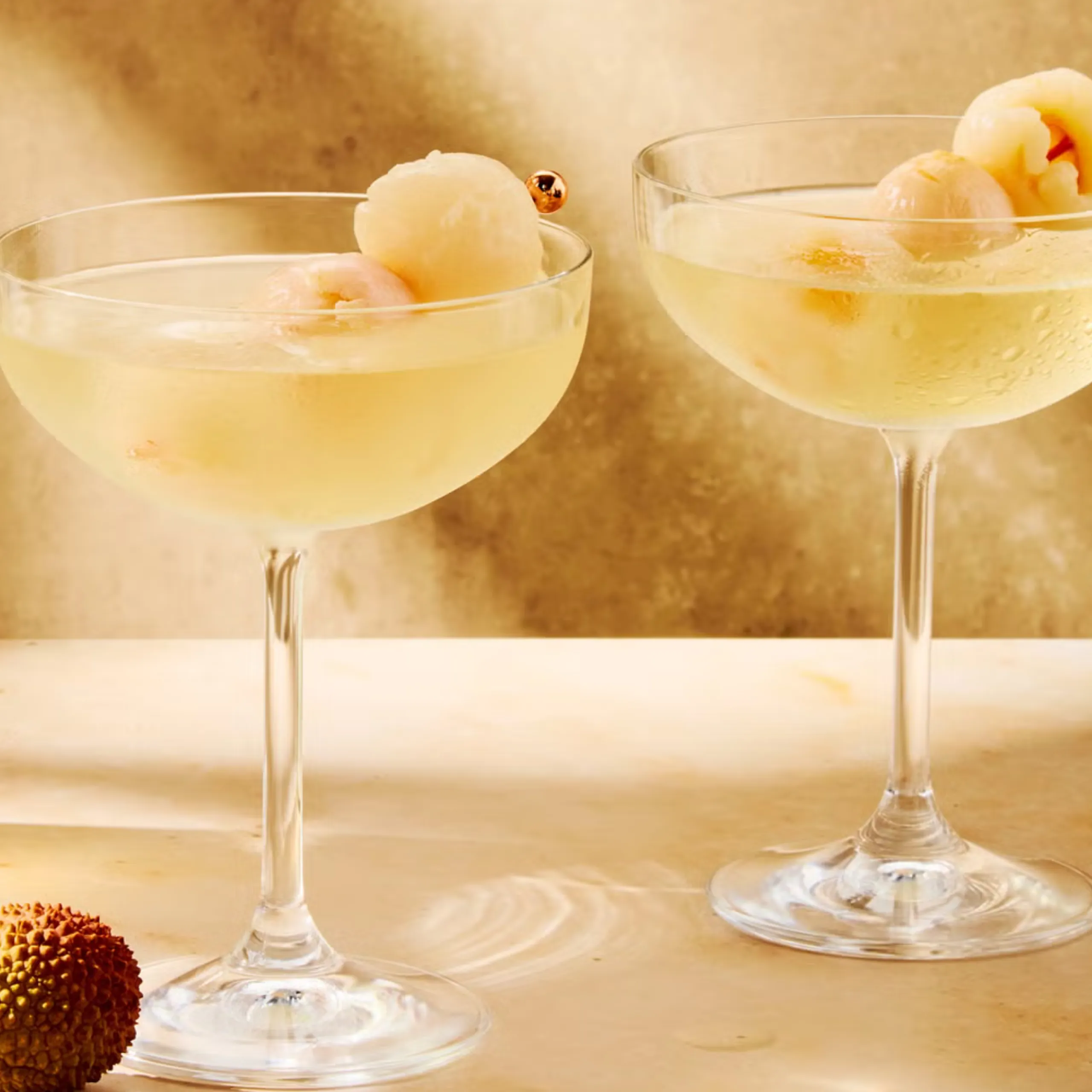 Two champagne coupes with pale cocktails and round white garnishes, a chocolate-dusted treat on the left foreground.