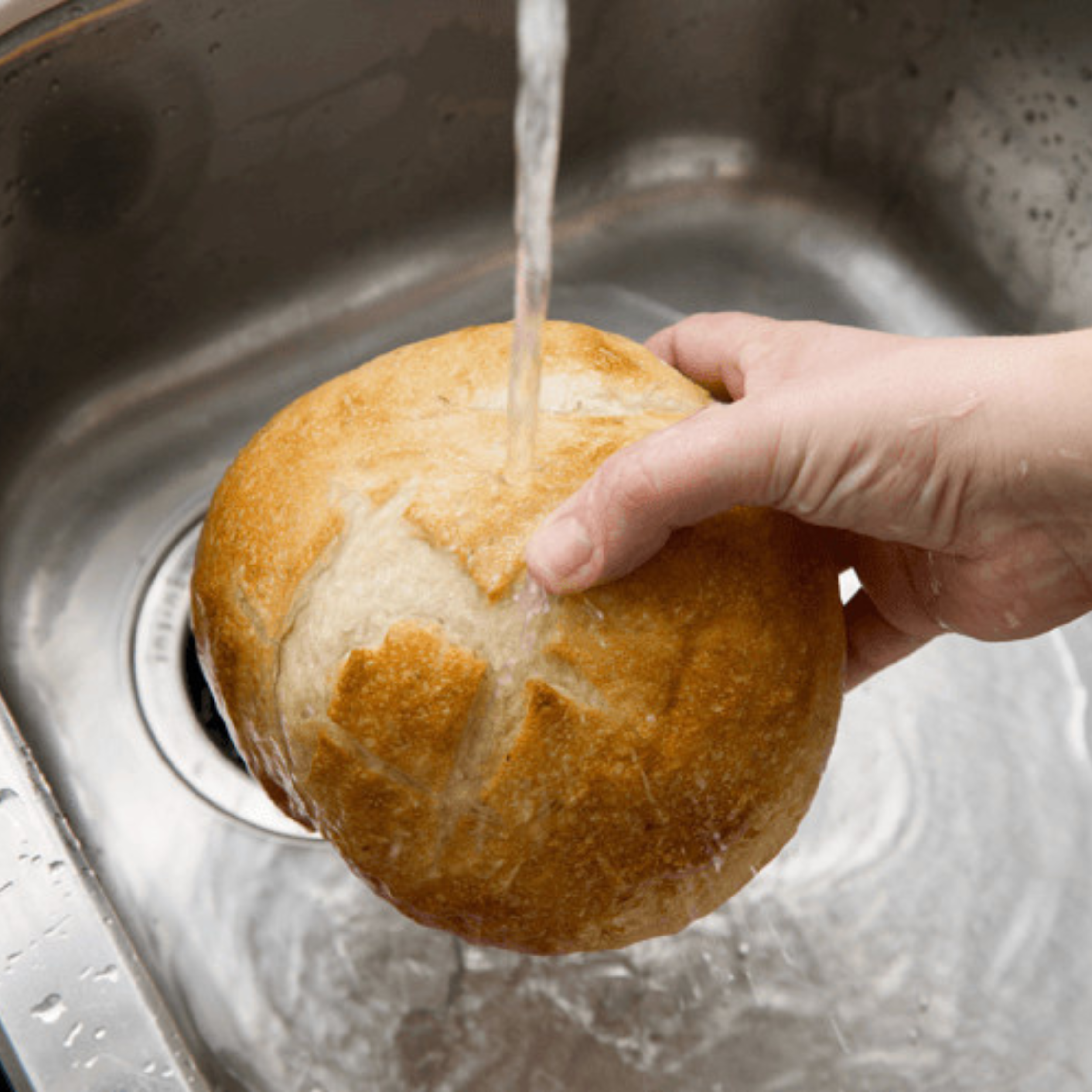 Close-up of a crusty bread roll being held by a hand as water runs from a faucet onto it over a metal kitchen scale in the background.