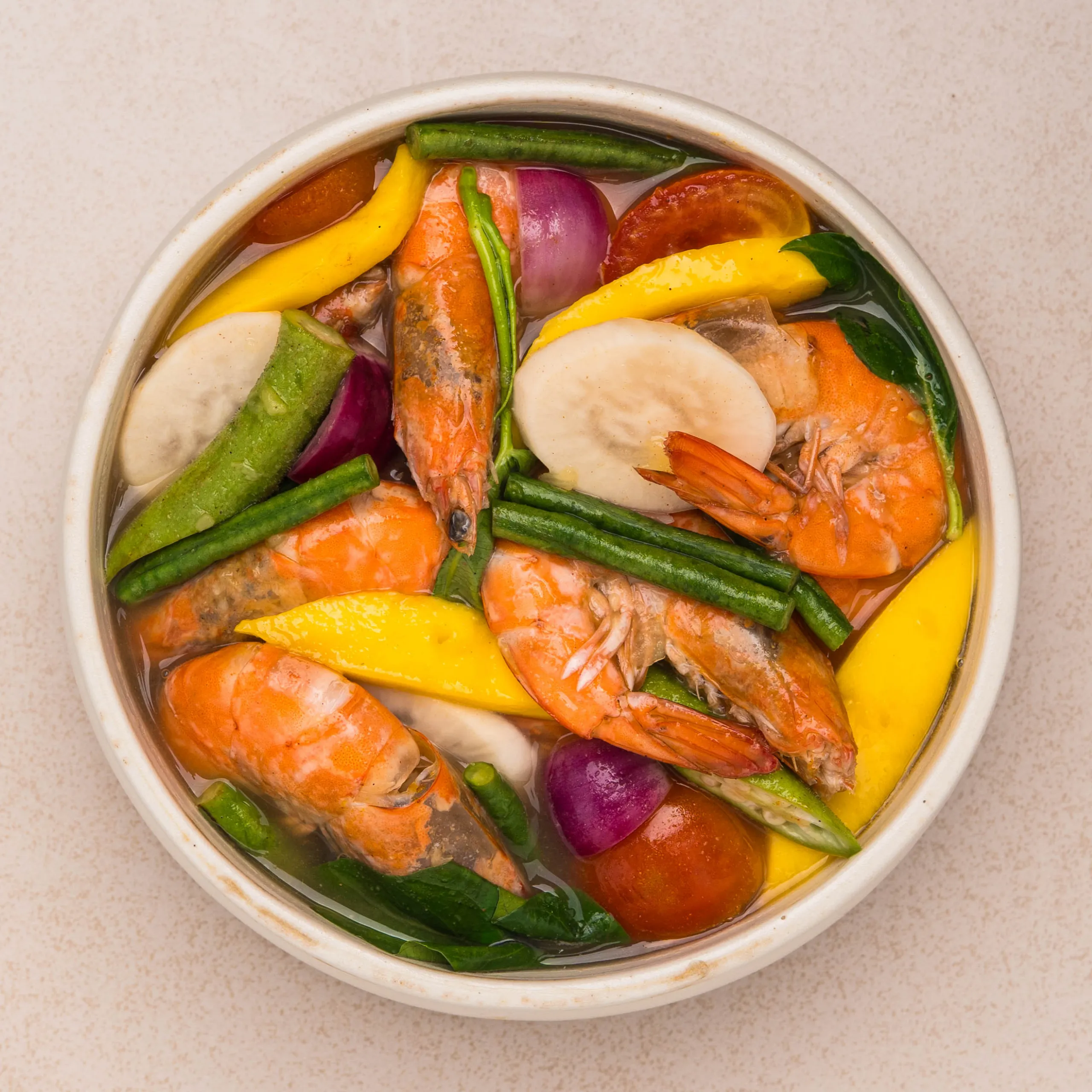 Top-down view of a pot filled with shrimp, yellow pepper strips, green beans, red onion, tomato slices, and greens in broth.