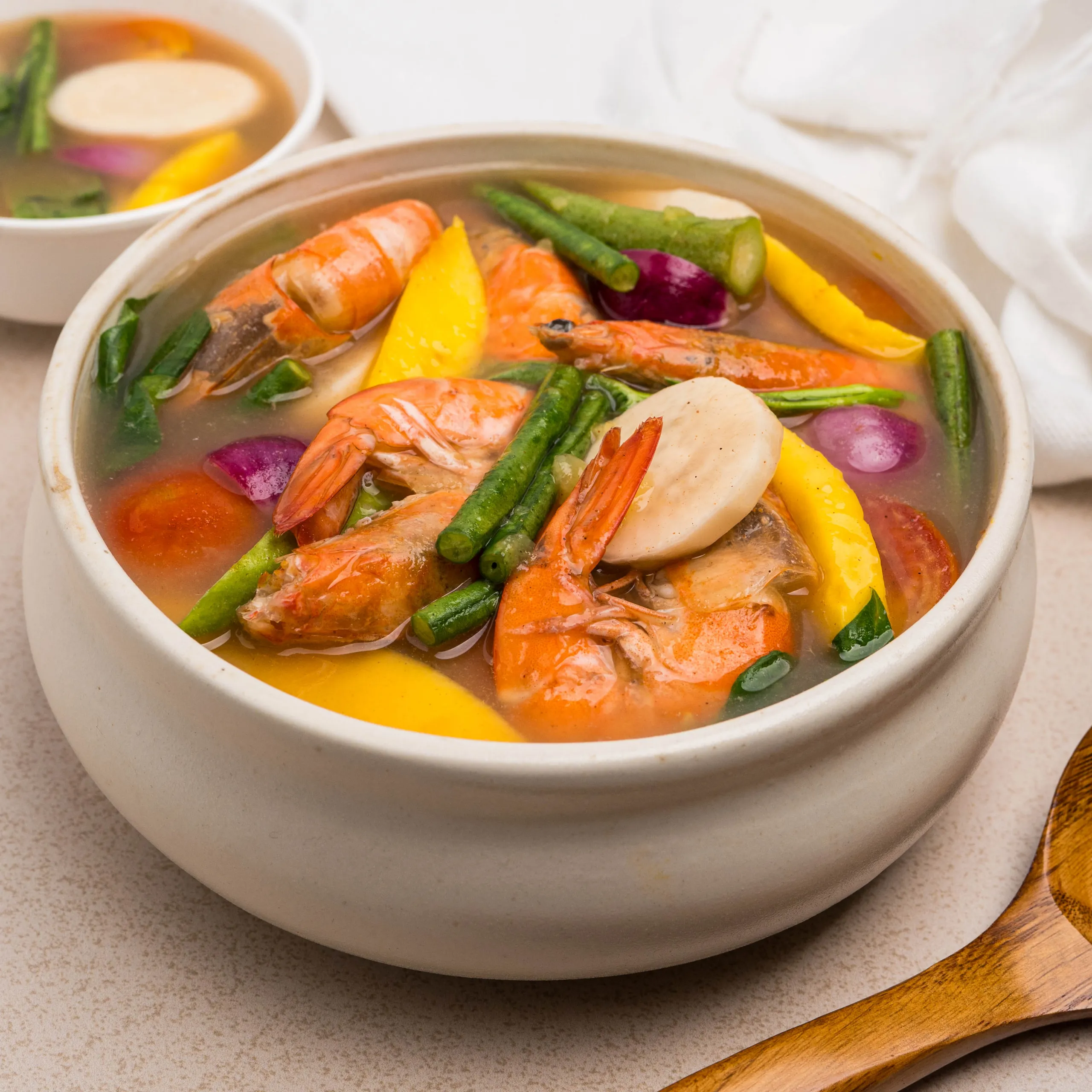 Close-up of a white bowl of shrimp soup with colorful vegetables (shrimp, green beans, yellow pepper, red onion) on a light countertop, with another bowl in the background.