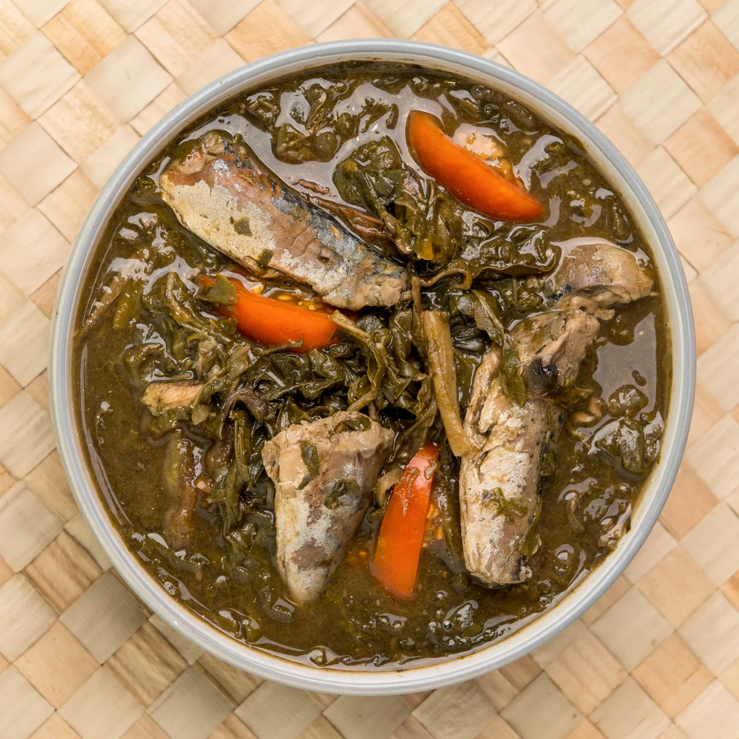 Top-down view of a pot filled with braised greens, chunks of meat, and orange carrot pieces in dark gravy on a woven mat.