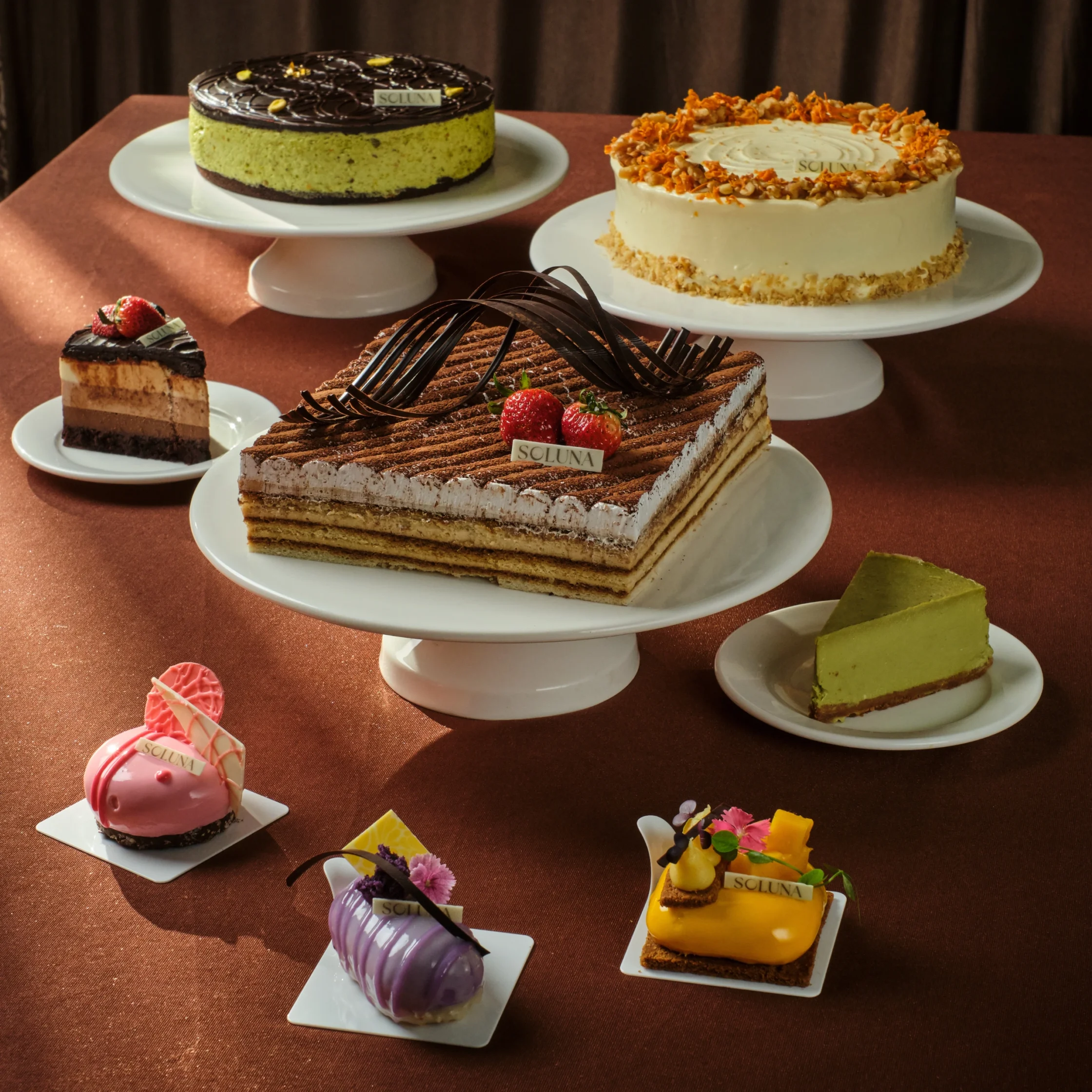 Assorted desserts on white stands: chocolate-glazed round cake, nut-crusted vanilla cake, and several small pastries on a brown table.