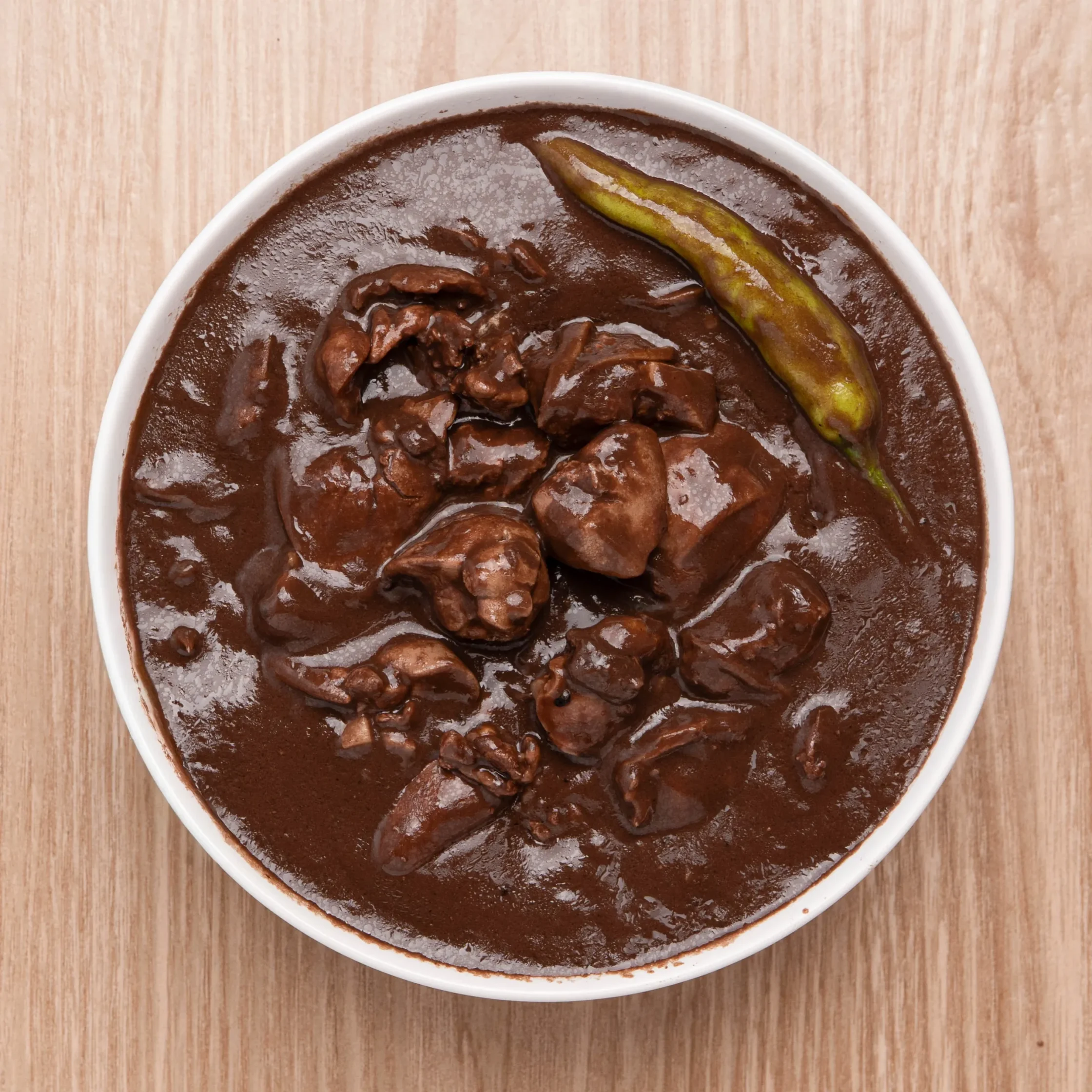 White bowl filled with thick dark chocolate mole sauce and chunks of meat, topped with a grilled plantain slice on a wooden table.