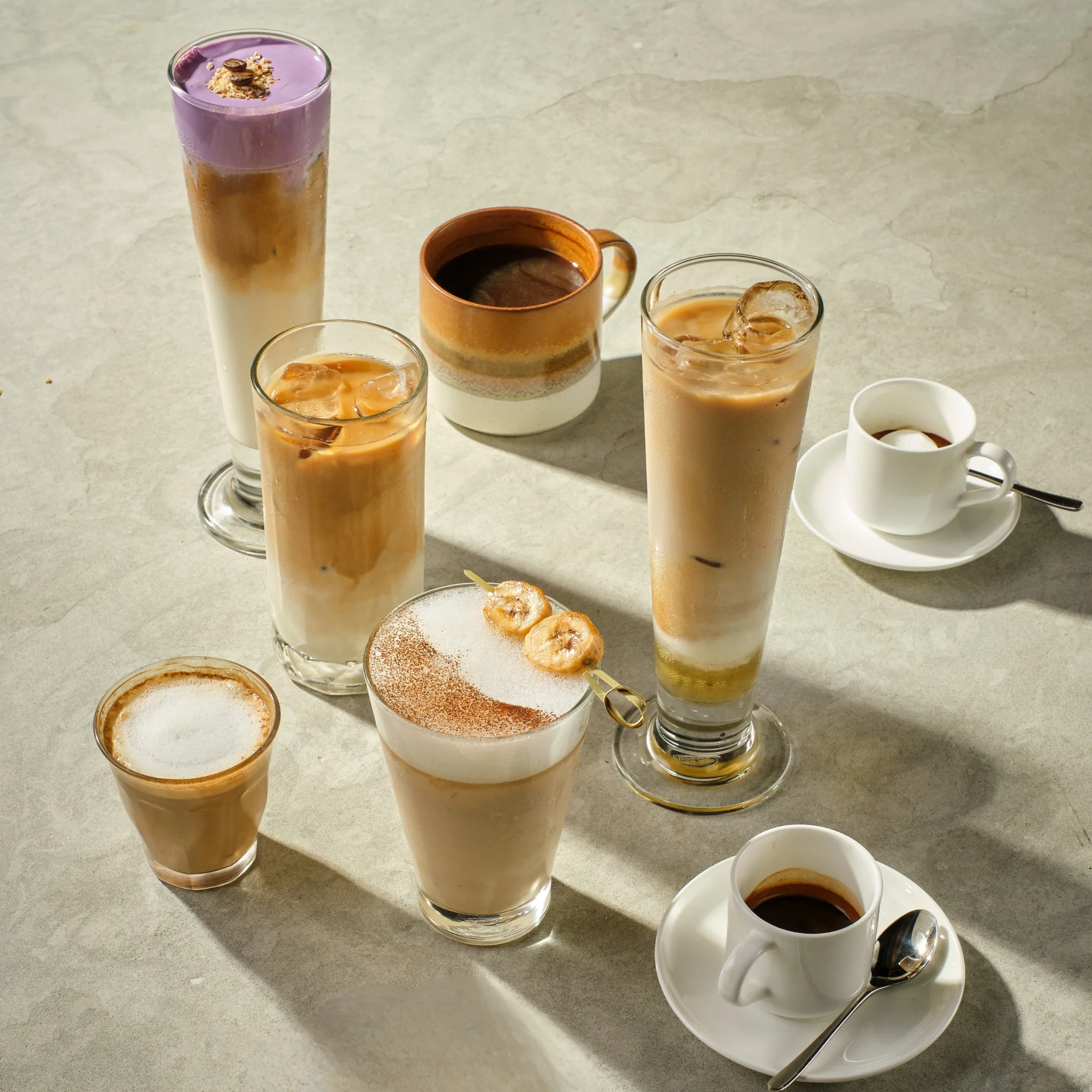 Tall glass of purple smoothie with granola topping, surrounded by layered iced coffee drinks on a light table.