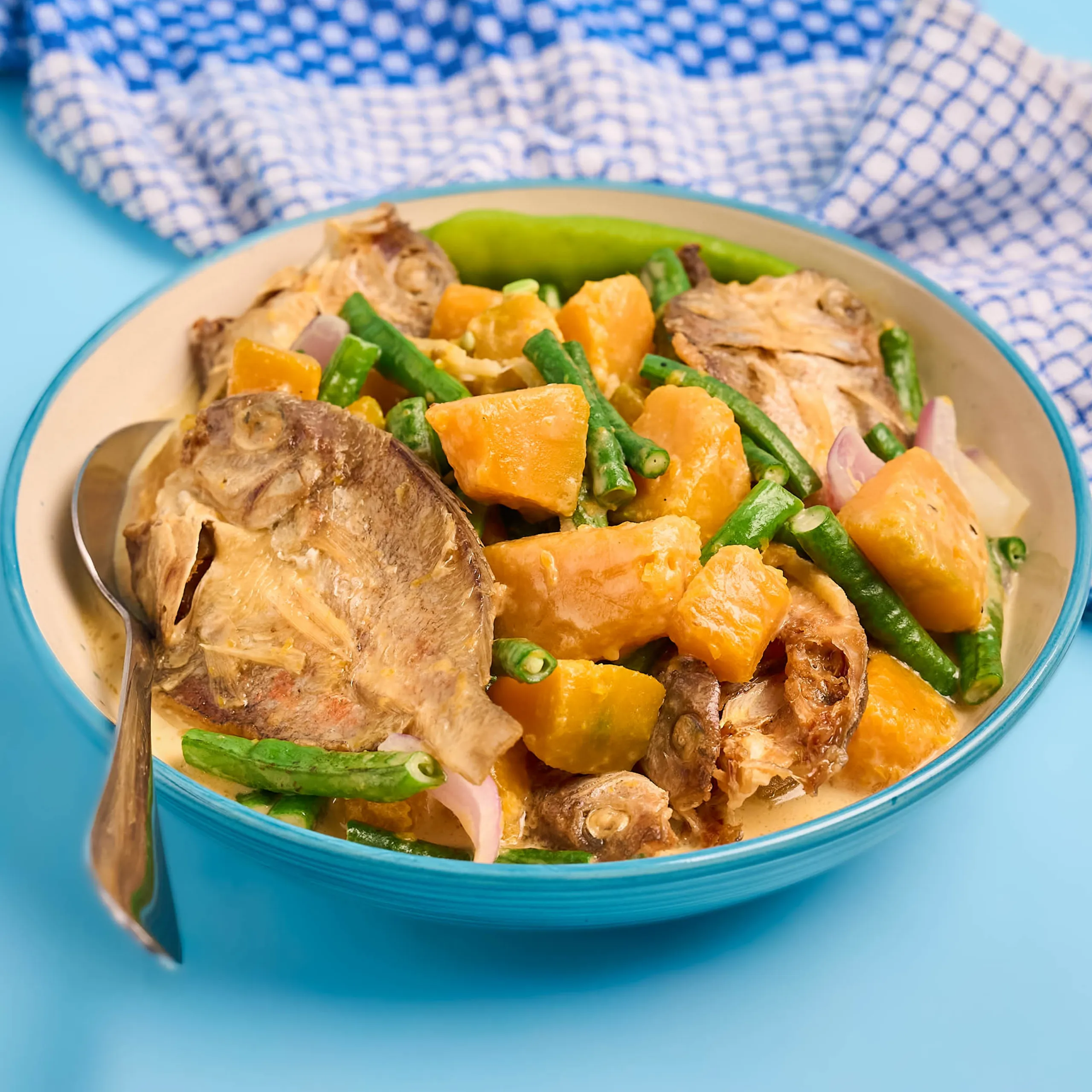 Bowl of cooked meat with orange cubed vegetables and green beans on a blue table, with a spoon resting inside the dish.