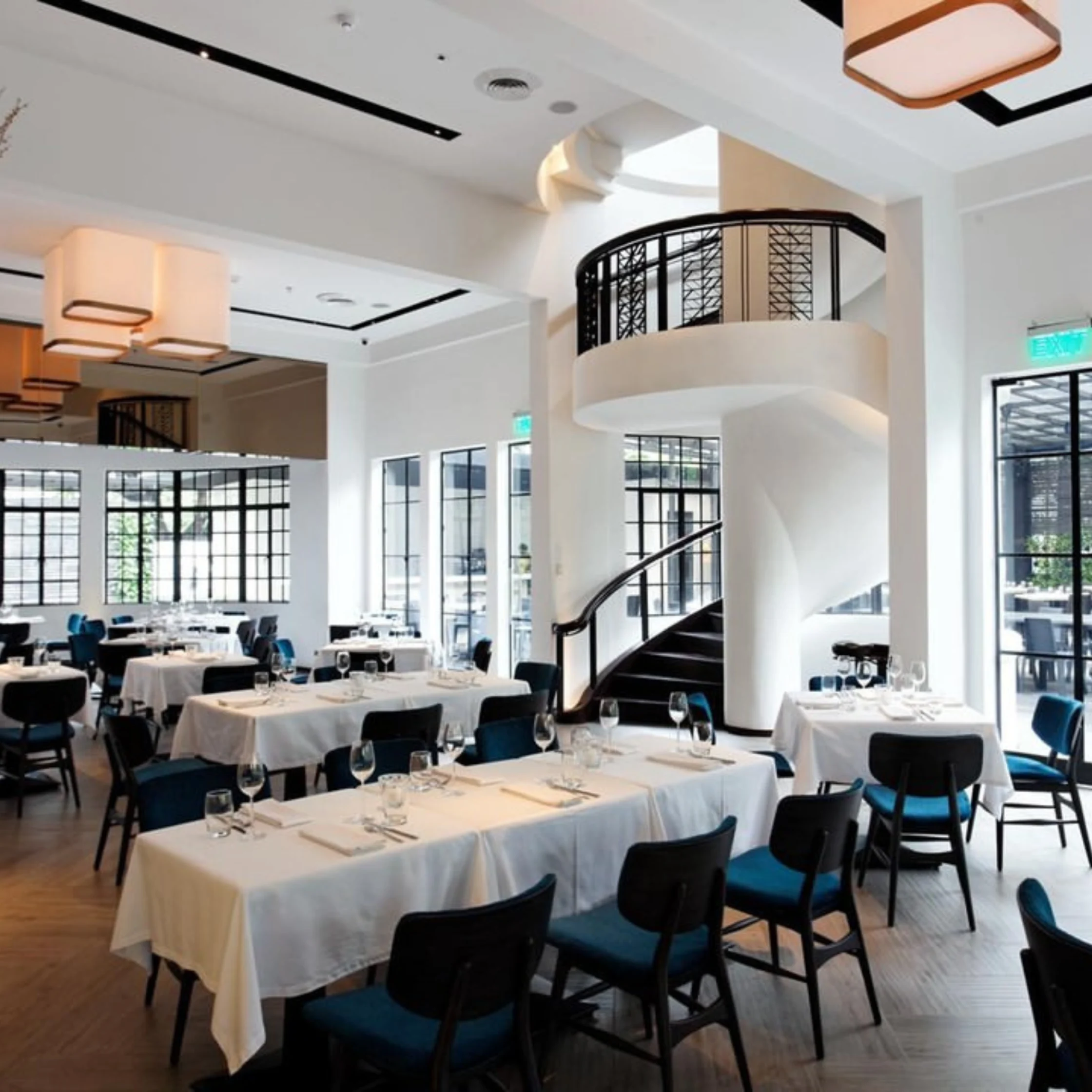 Banquet hall with white-clothed tables and blue chairs in a modern restaurant interior, curved staircase in the background.