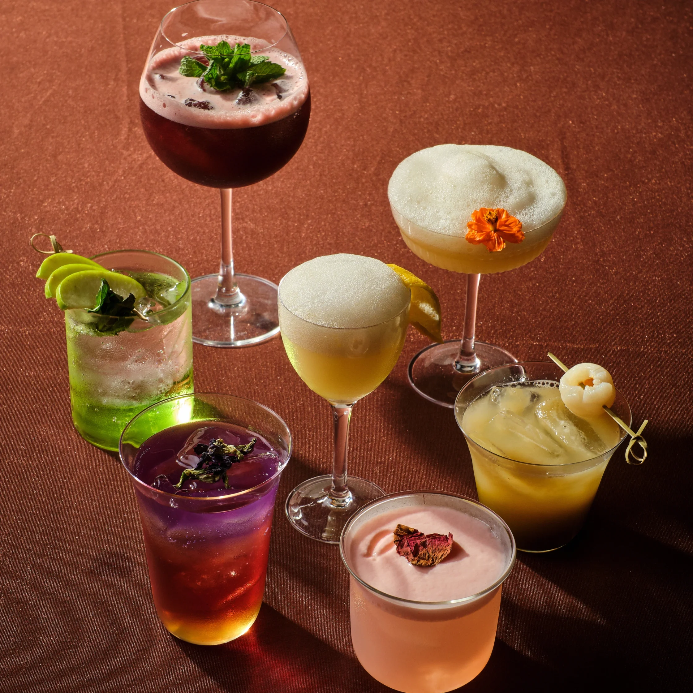 Collection of colorful cocktails in stemmed glasses on a textured copper background; includes a dark red drink with mint garnish, a pale pink foamy cocktail, a bright green mojito with lime wedges, a pale yellow drink with an orange flower, and a frothy pale beverage with a lemon twist.