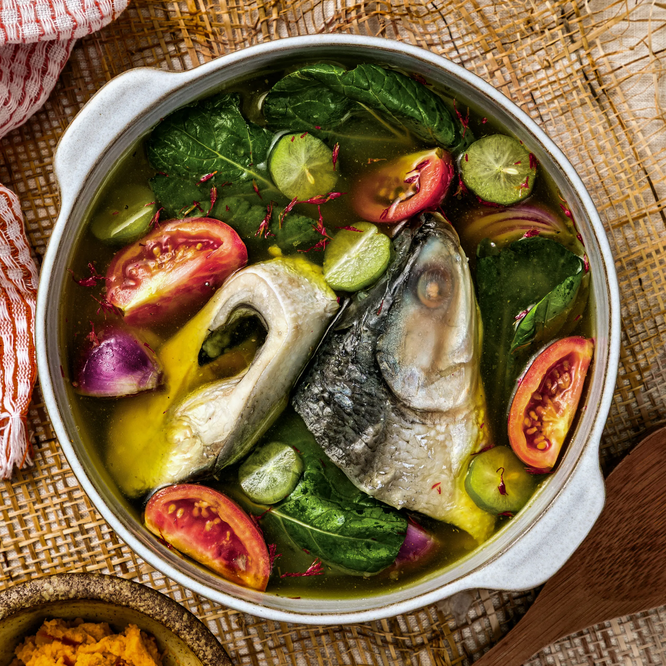 Pot of fish soup with leafy greens, tomato wedges, lime slices, and red onion in broth on a woven mat.