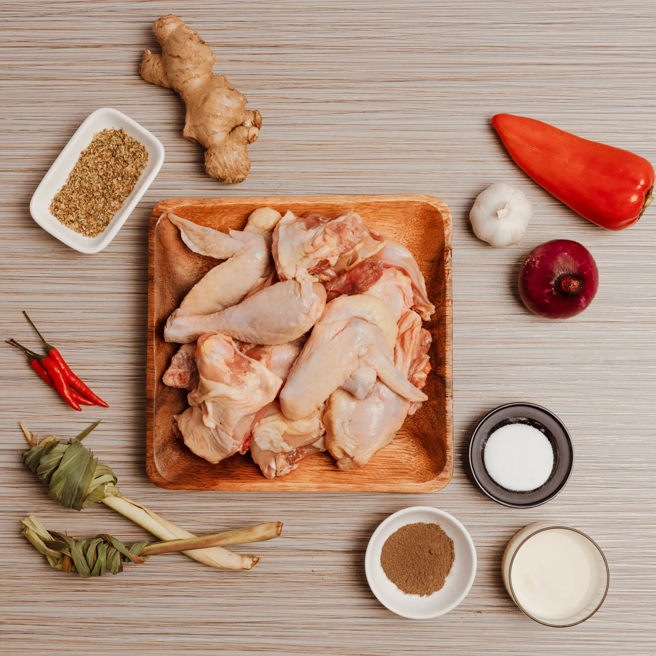 Raw chicken pieces on a wooden cutting board with bowls of spices and assorted aromatics nearby (garlic, lemongrass, red pepper).