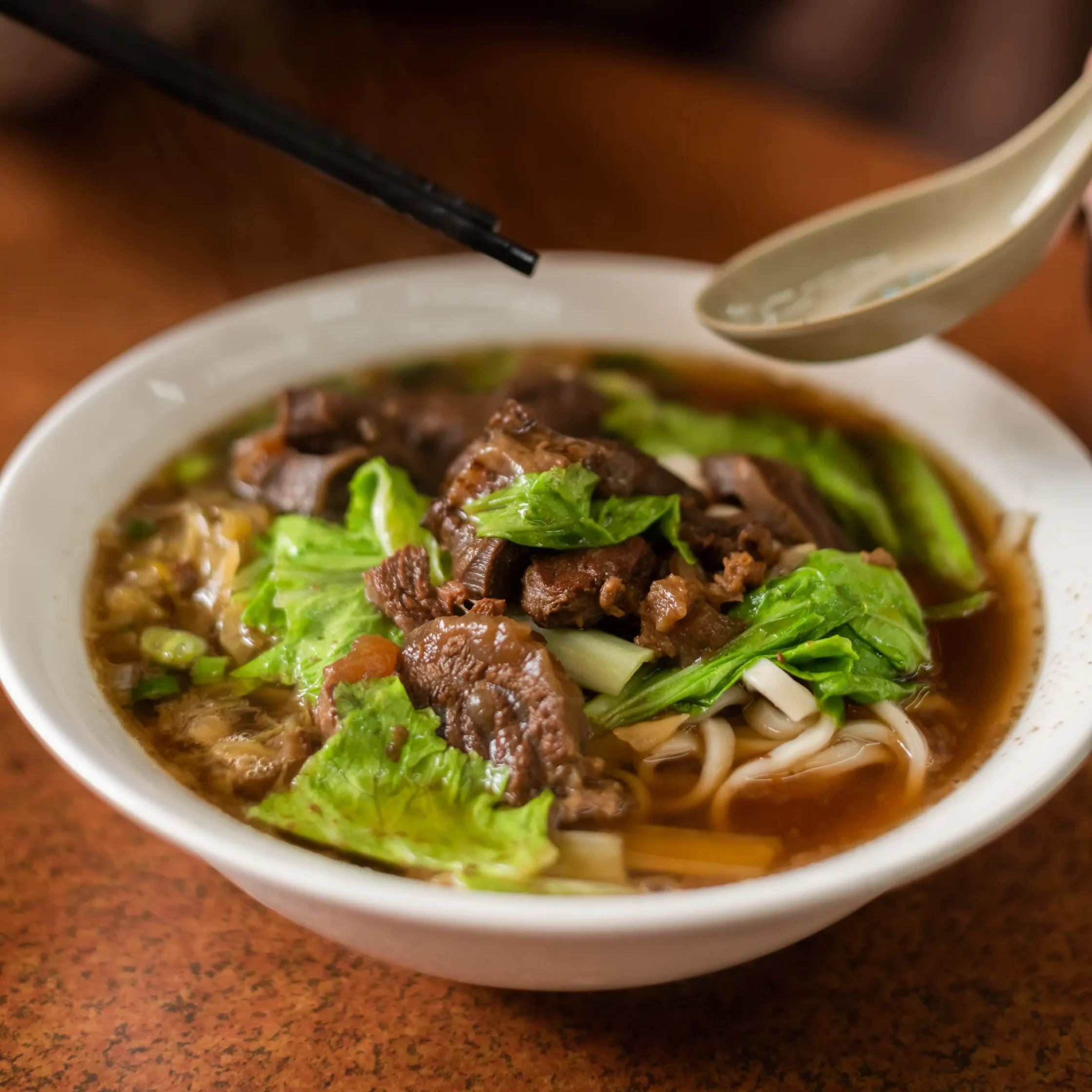 Bowl of beef noodle soup with greens, viewed from above; black chopsticks hovering above the noodles.