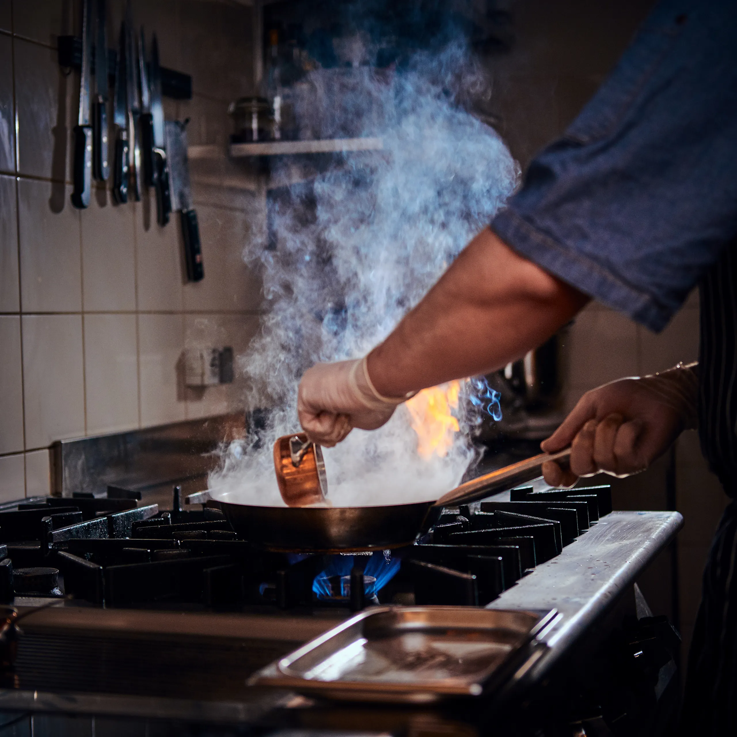 Hands stirring a copper pan on a gas stove with visible blue flames and rising steam in a kitchen setting