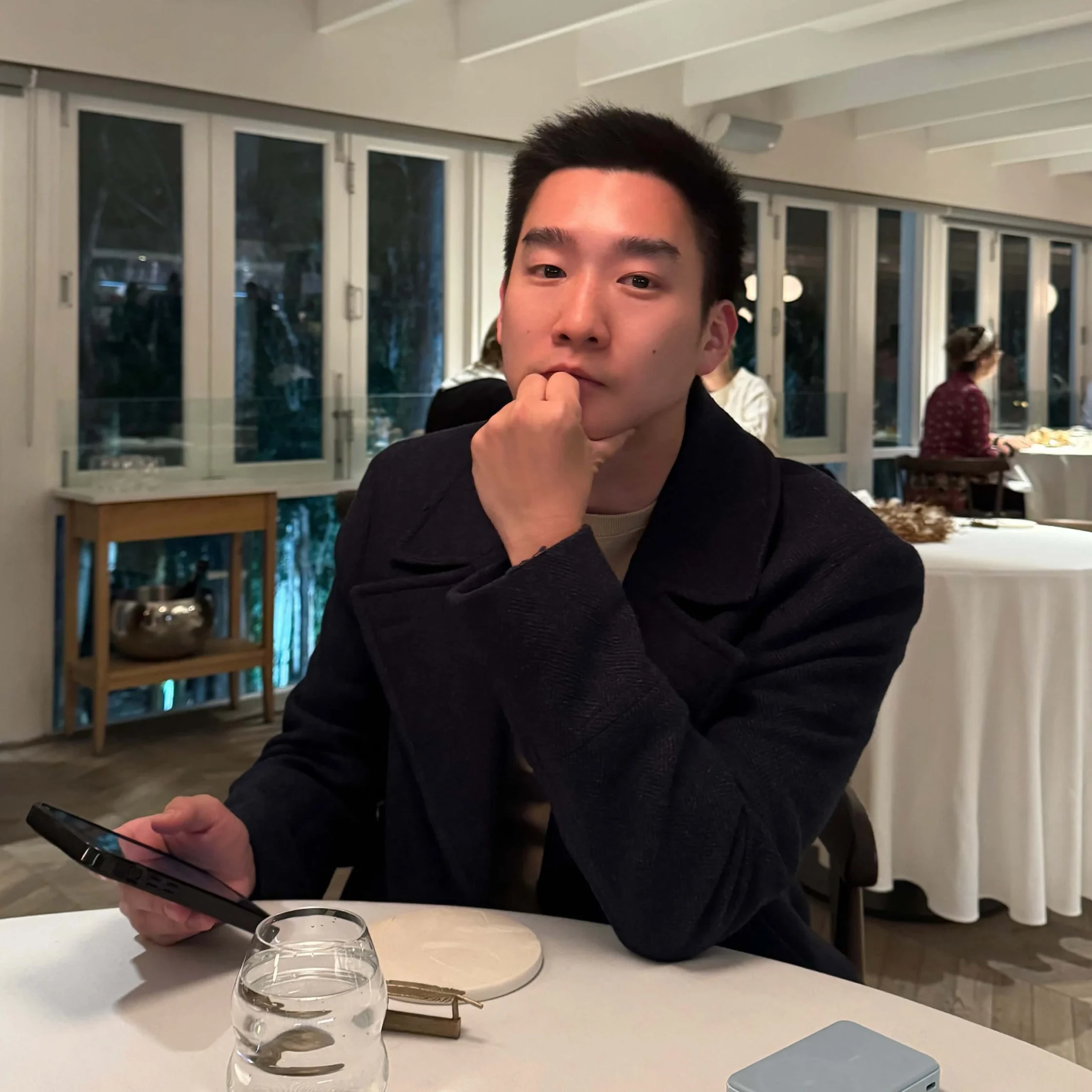 Man seated at a white table in a restaurant, looking at his smartphone with his elbow resting on the table.