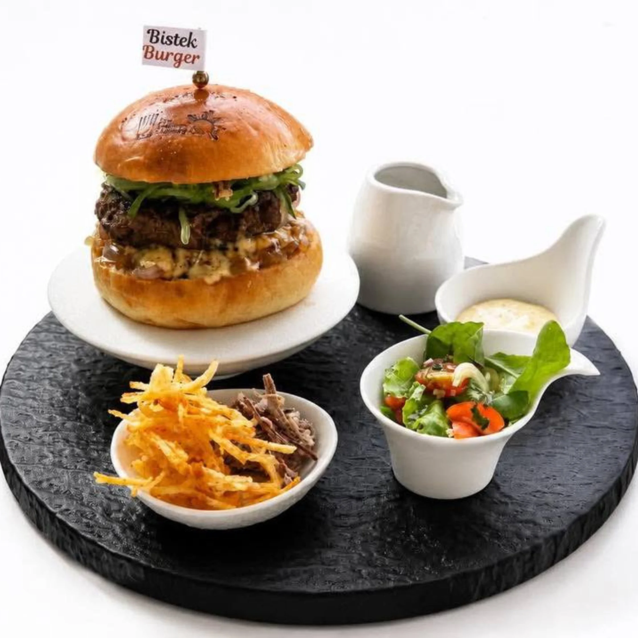 Burger served on a black slate platter with sides: a small salad cup, crispy onion chips, and a white sauce jug, topped with a small flag reading 'Bistek Burger Best Chef of the Year chef John Carlo Palacol Entry at PCC