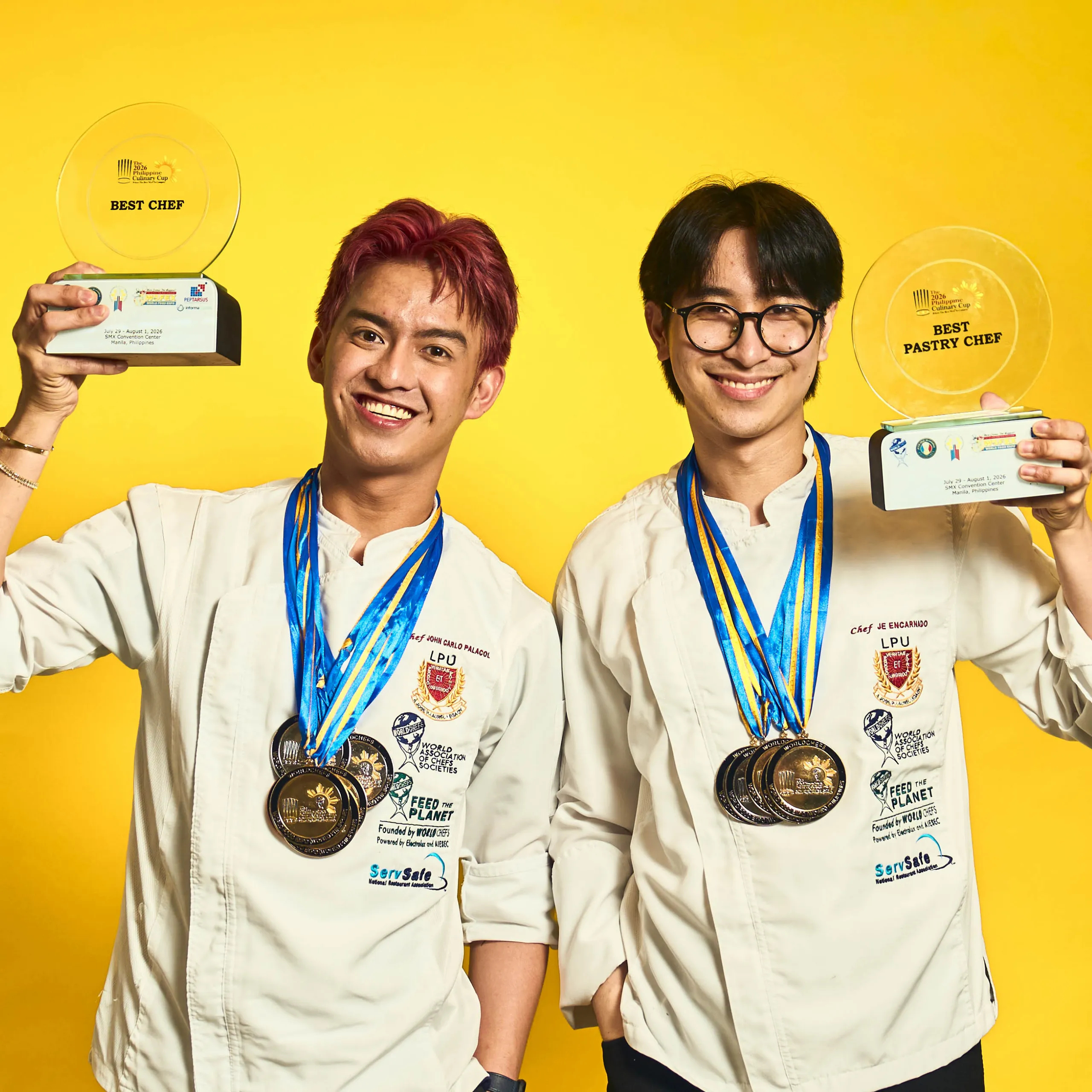Two chefs in white coats wear blue ribbons and smile, holding large trophy plaques and many medals against a bright yellow background for a celebratory photo. Best Chef of the Year chef John Carlo Palacol and Best Pastry Chef of the Year Jeriel Encarnado of LPU Laguna