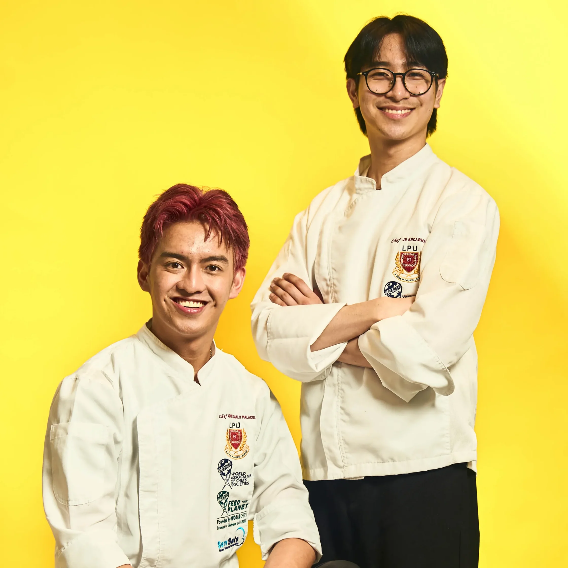 Two chefs in white coats wear blue ribbons and smile, holding large trophy plaques and many medals against a bright yellow background for a celebratory photo. Best Chef of the Year chef John Carlo Palacol and Best Pastry Chef of the Year Jeriel Encarnado of LPU Laguna