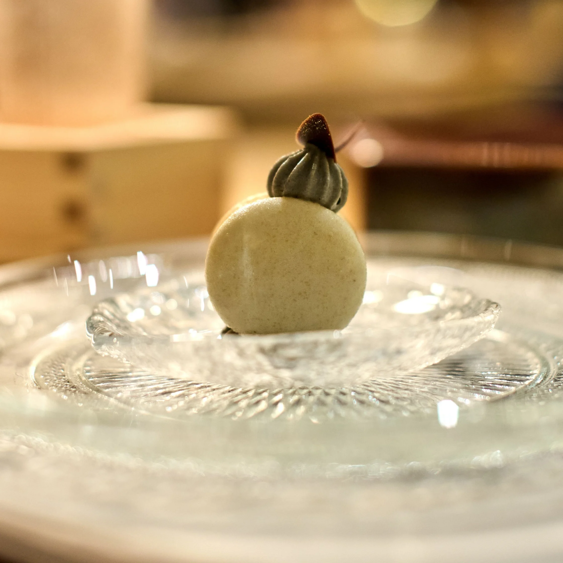 Close-up of a cupcake with pale frosting and a chocolate-topped peak, on a glass plate with a blurred background.