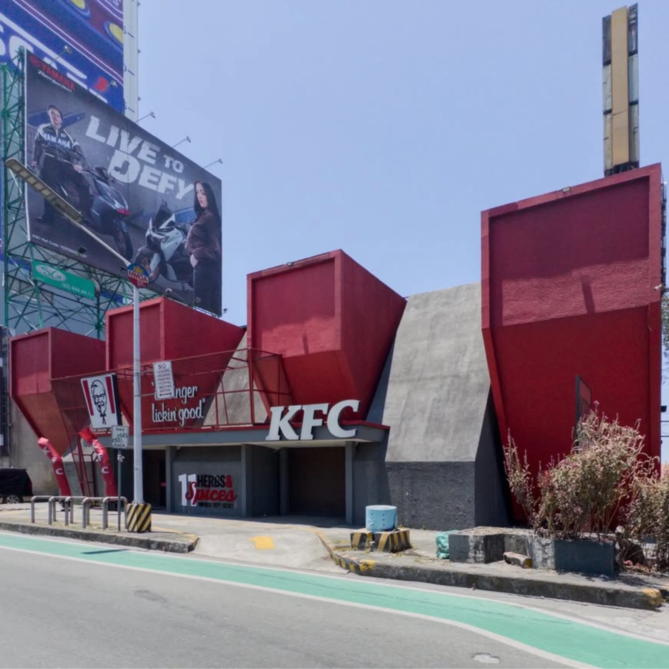 Exterior view of a KFC restaurant with red building and large advertising billboards on the left against a clear blue sky. A bike lane with green pavement runs along the road.