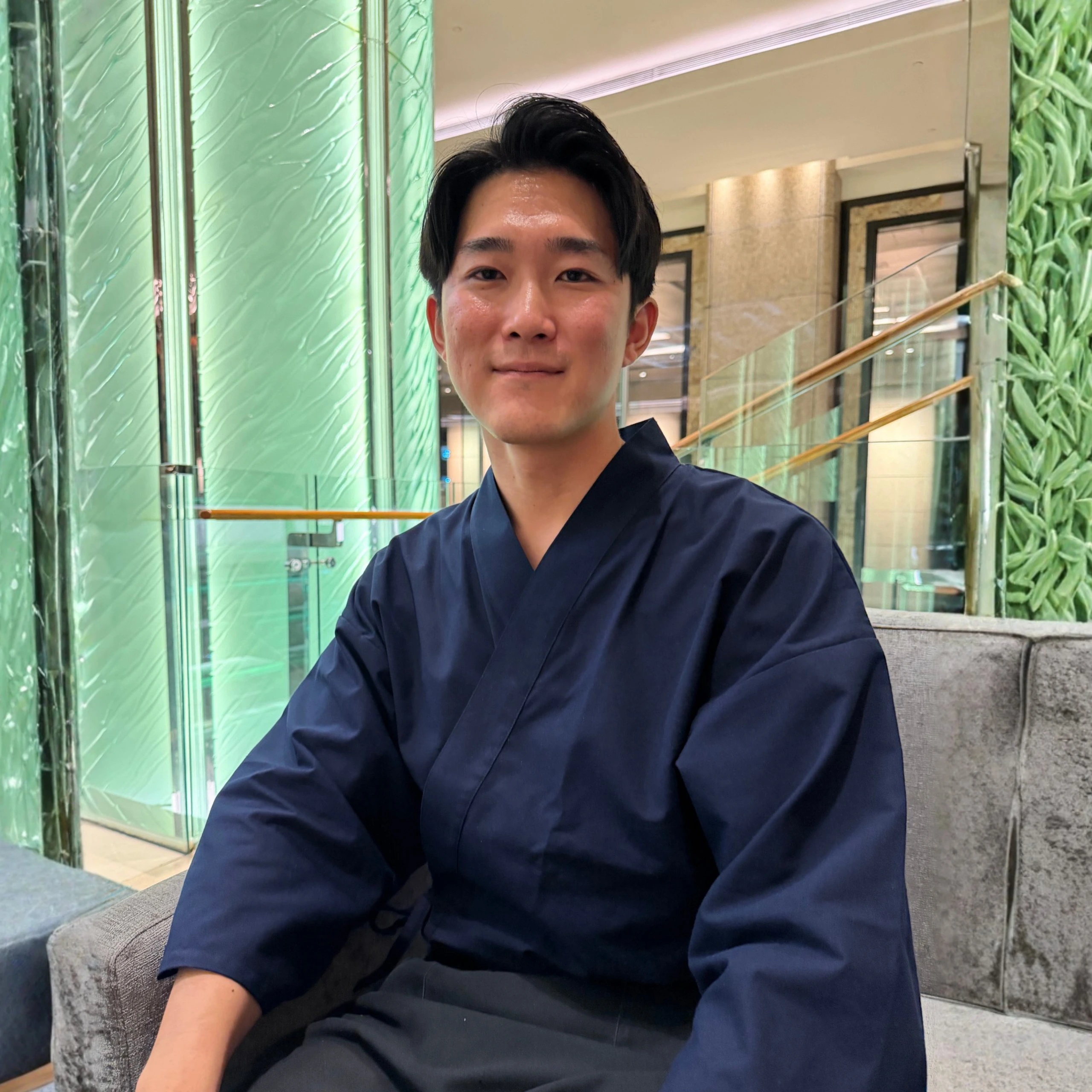 Man in dark blue kimono smiling and sitting on a couch in a place with green interiors