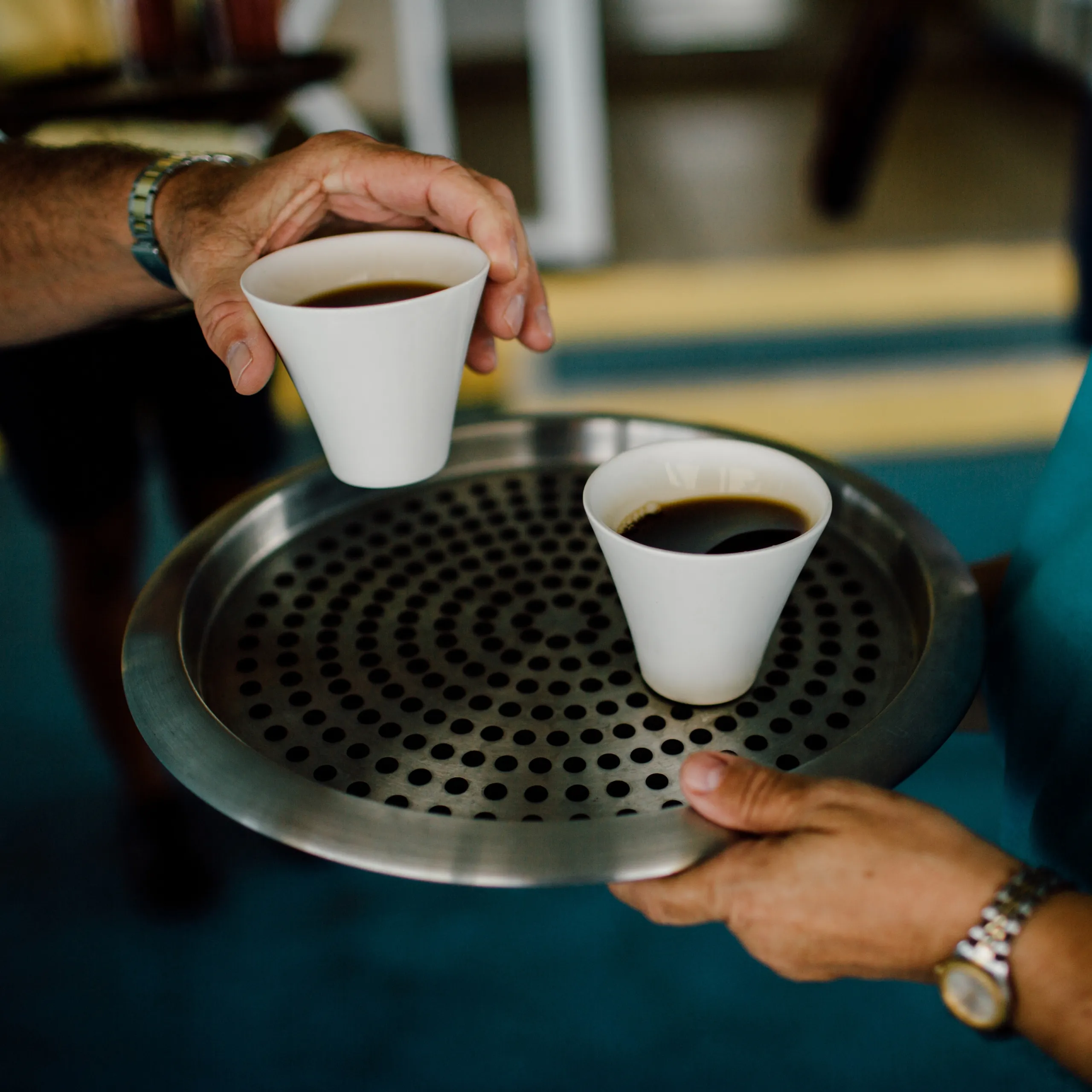 Hands hold two white paper coffee cups over a perforated metal tray; dark coffee fills the cups.