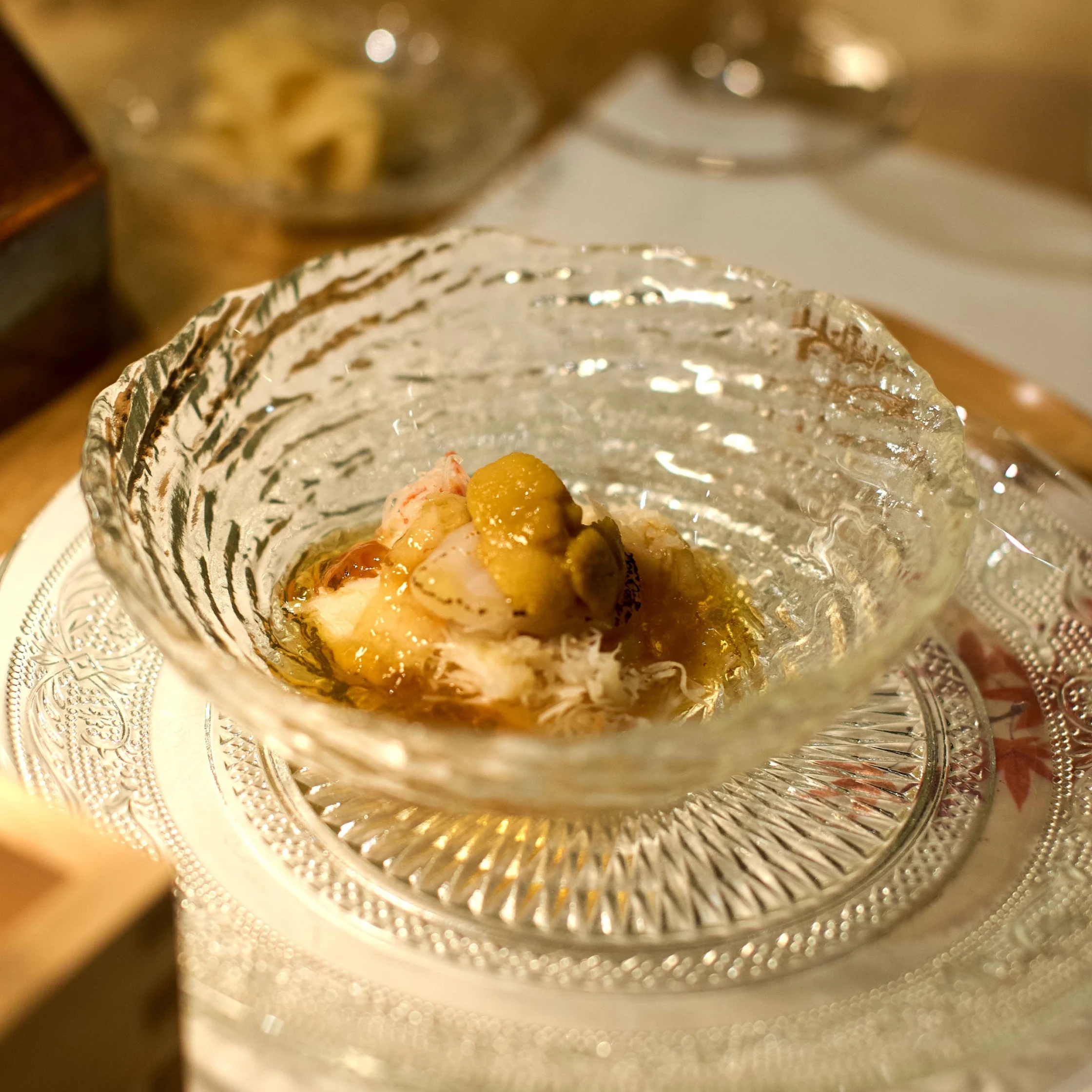 Close-up of a plated seafood dish in a decorative glass bowl with foil edge, small orange garnish on top.