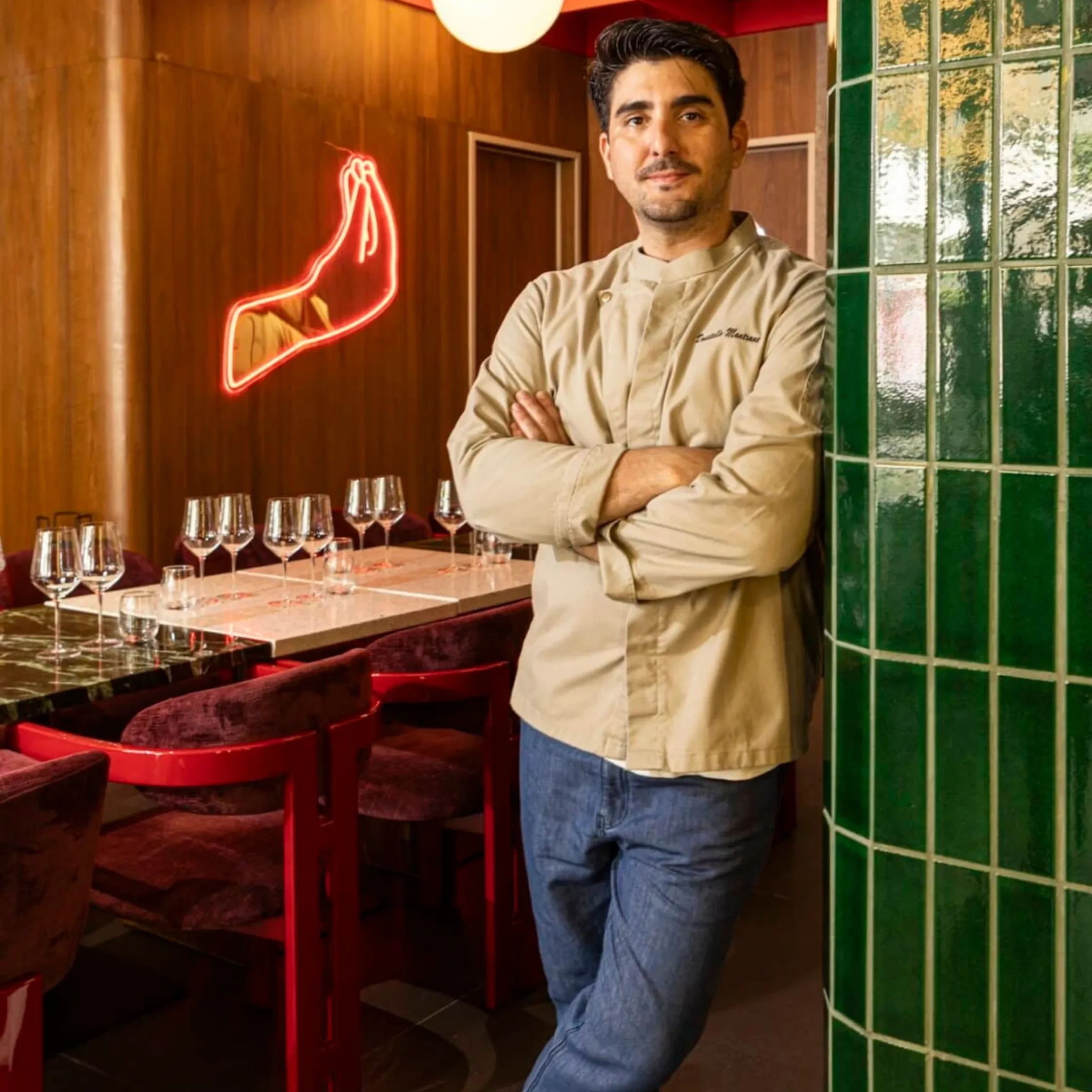 Chef in beige uniform stands with arms crossed in a stylish restaurant, neon sign glowing behind him.