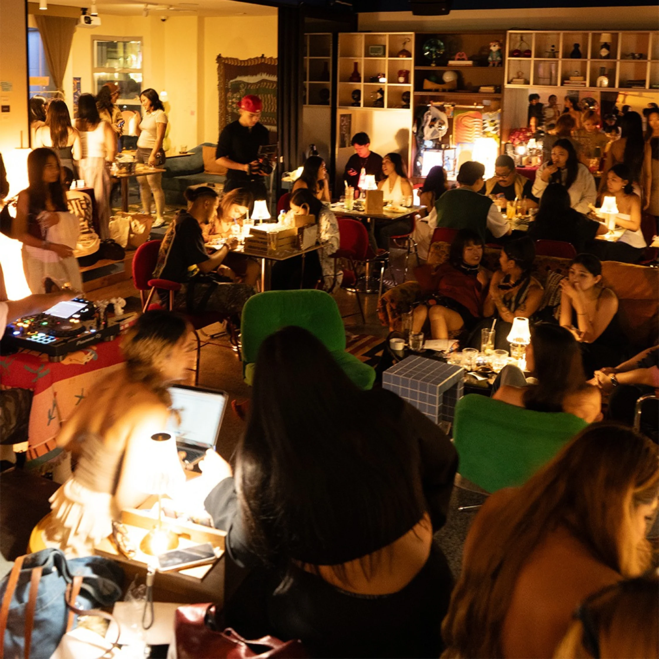 Crowded, warmly lit restaurant or party space with people eating and chatting at tables; a DJ mixer visible in the foreground.