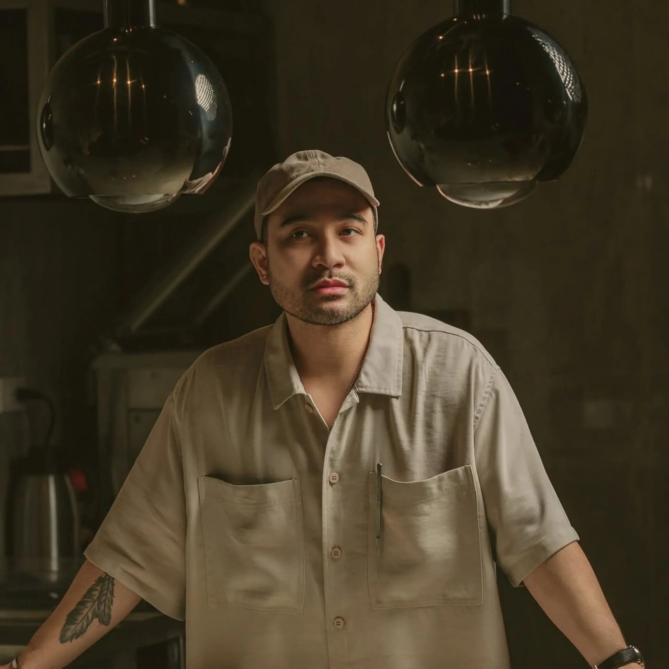 Man in a beige button-up shirt and cap stands indoors with his arms slightly extended; visible tattoo on his forearm in a dimly lit room with hanging globe lights behind him.