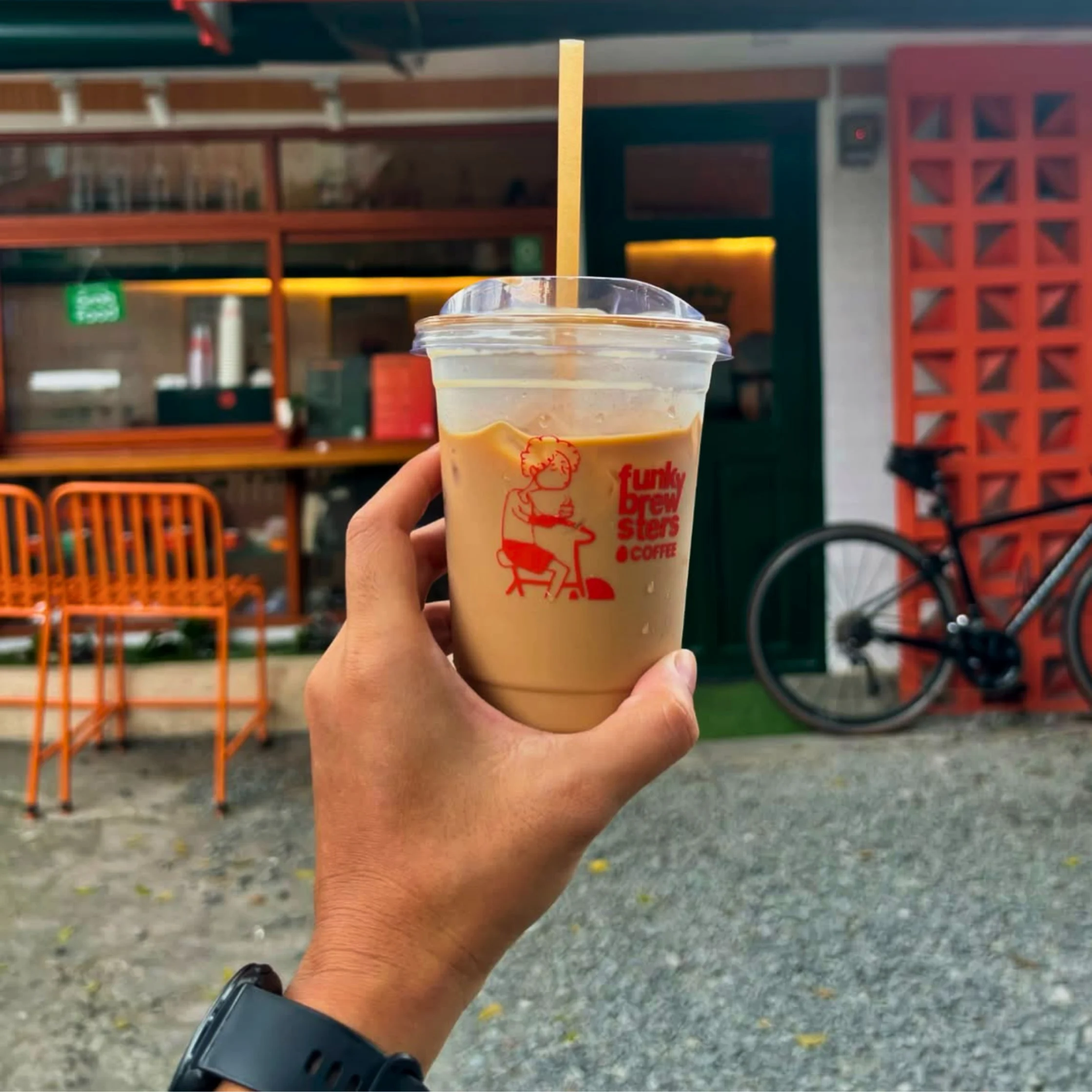 Hand holding an iced coffee cup with a straw in the foreground, at an outdoor cafe with orange chairs and a brick red lattice wall in the background BF Homes Parañaque