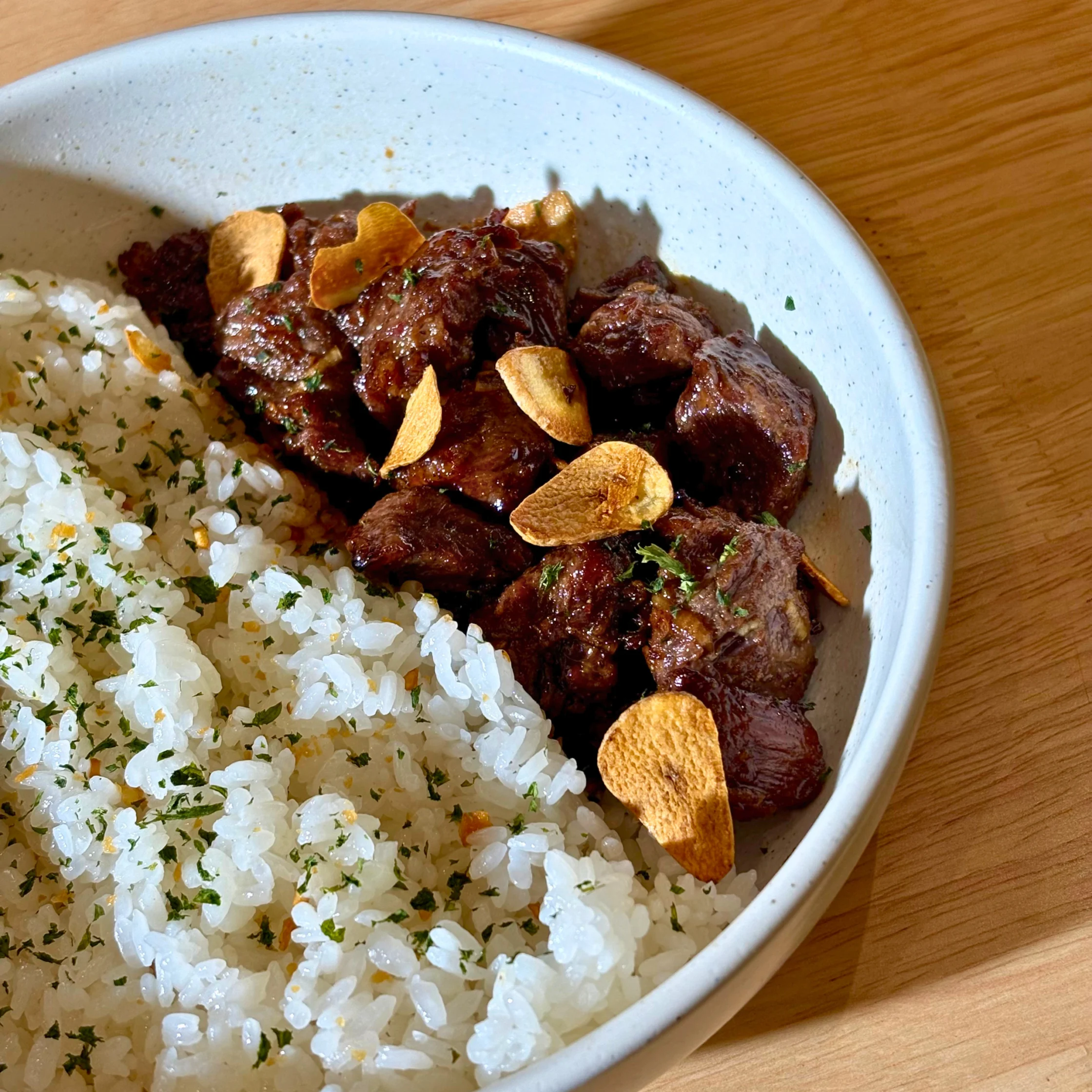 Beef slices with crispy garlic chips served over herbed white rice on a light plate.