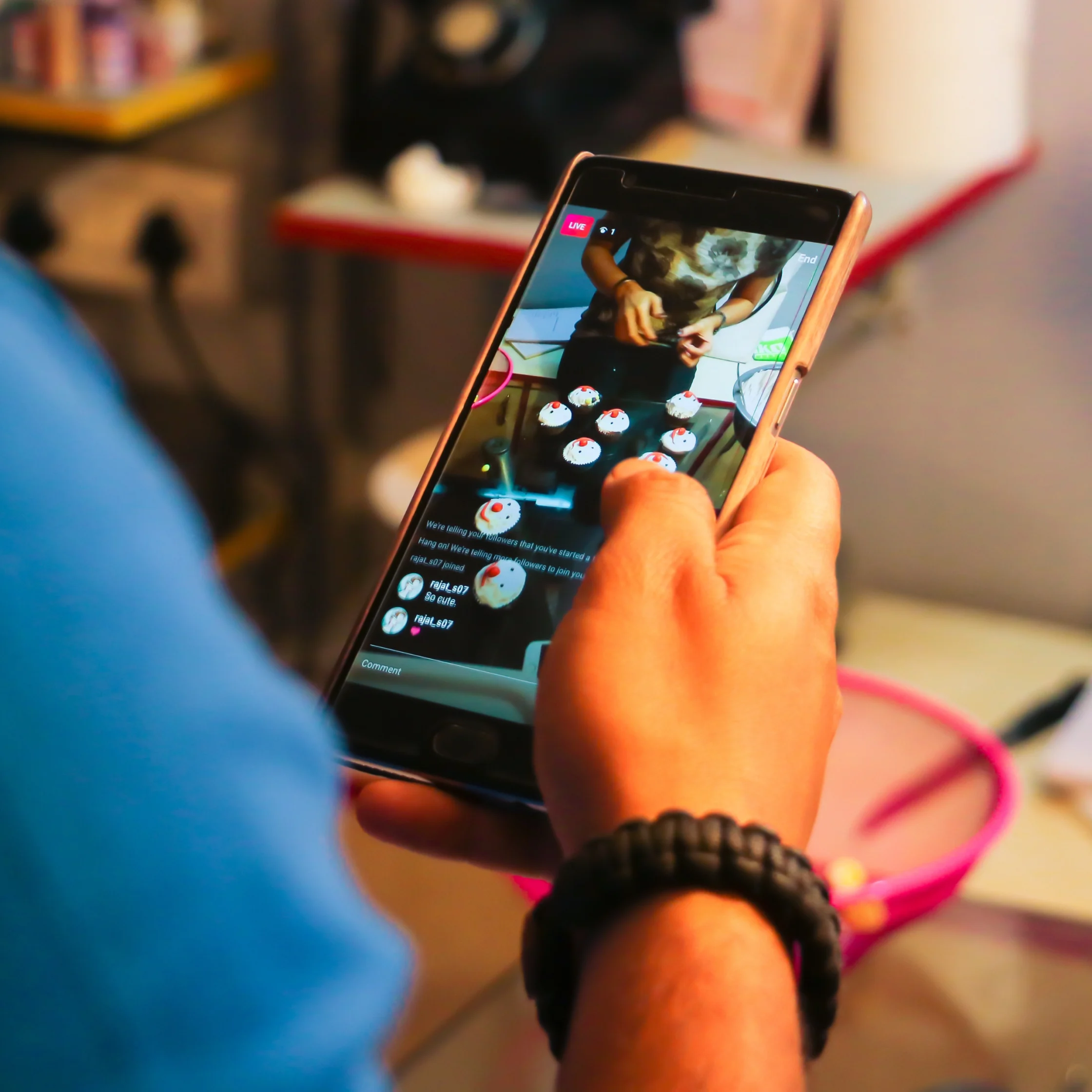 Person holding a smartphone showing a live video of a person decorating cupcakes at a table; scene viewed through the phone screen.