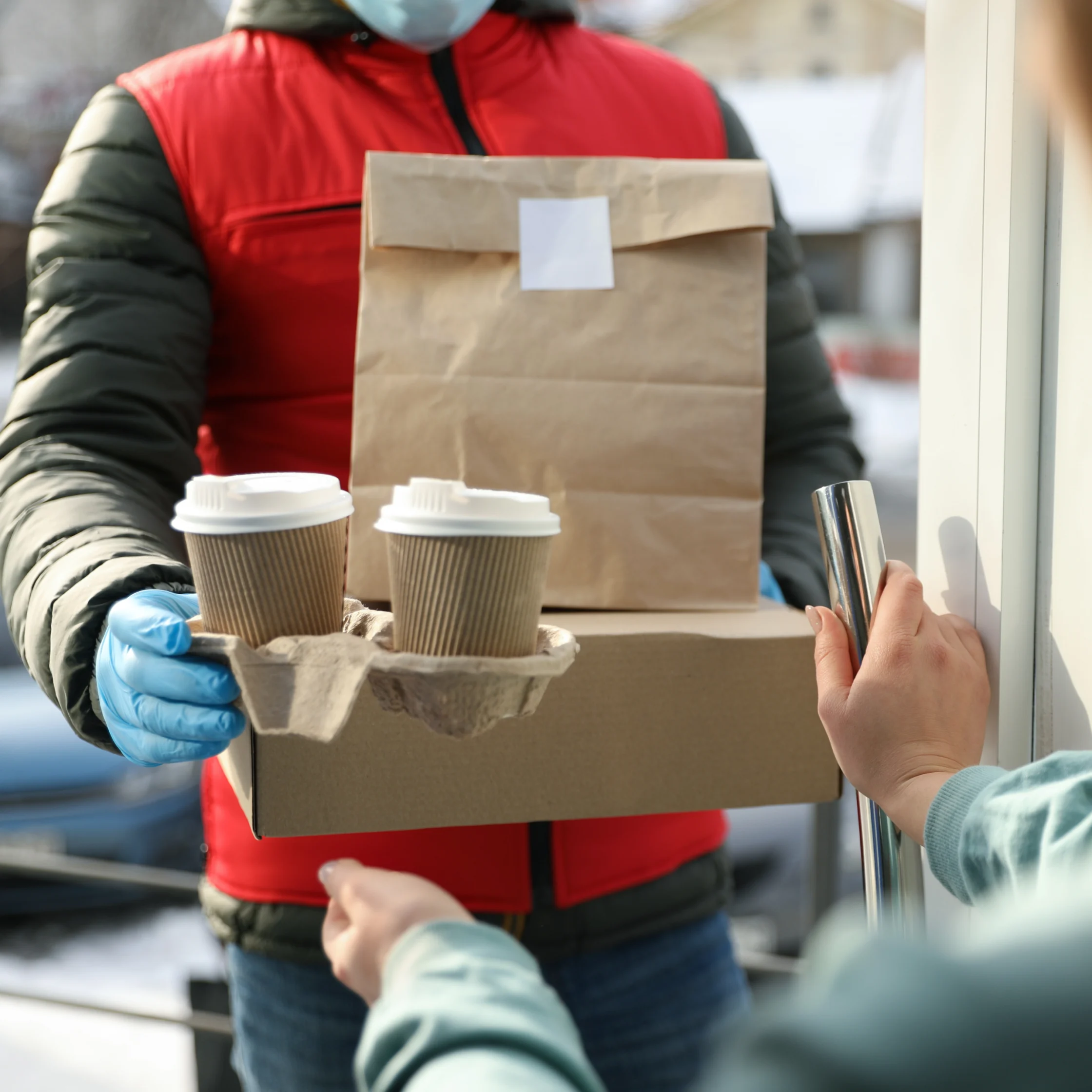 Delivery person in a red jacket hands a brown paper bag to a customer at a doorway, with two coffee cups in the foreground.