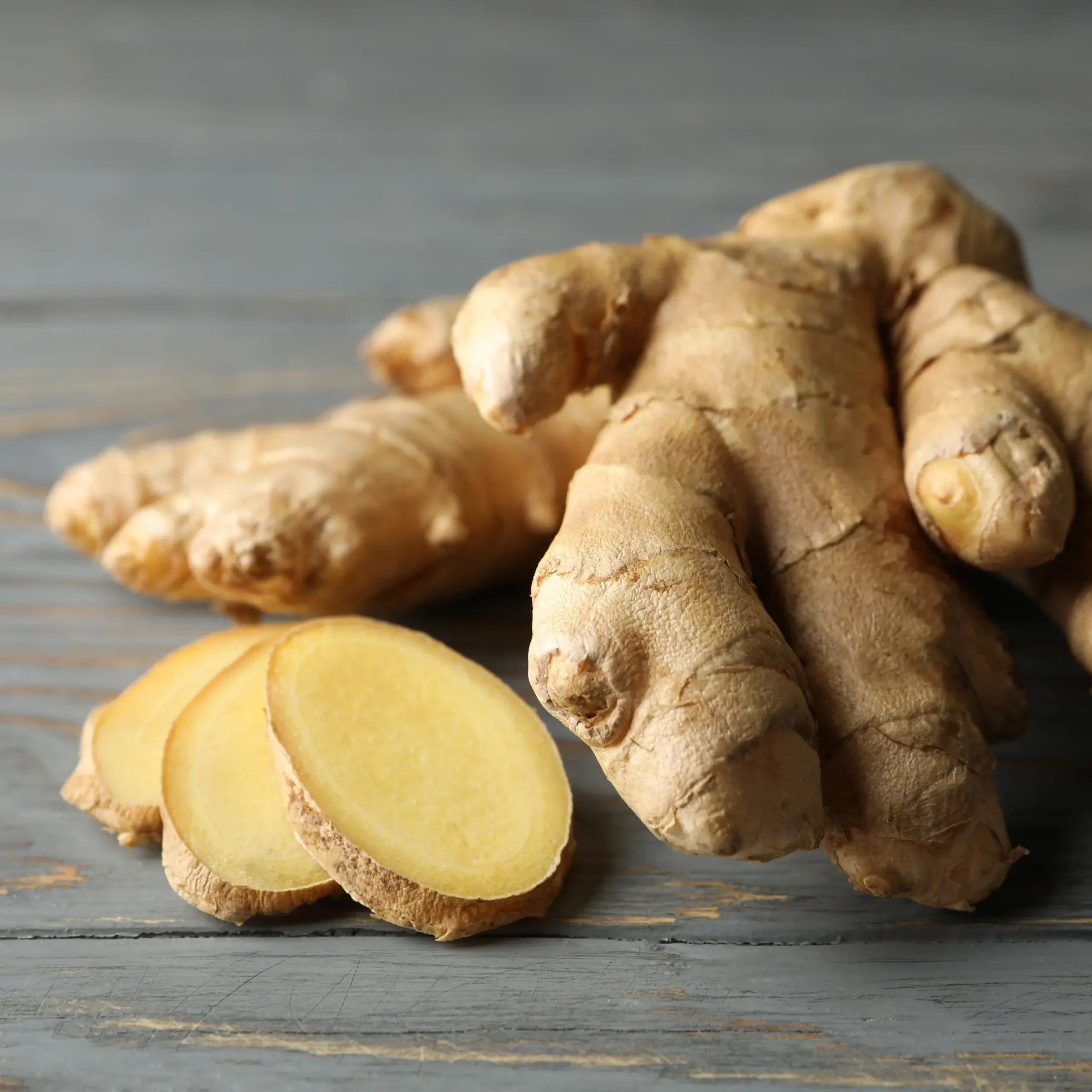 two pieces of unpeeled ginger next to three sliced pieces of giner on a grey wooden table