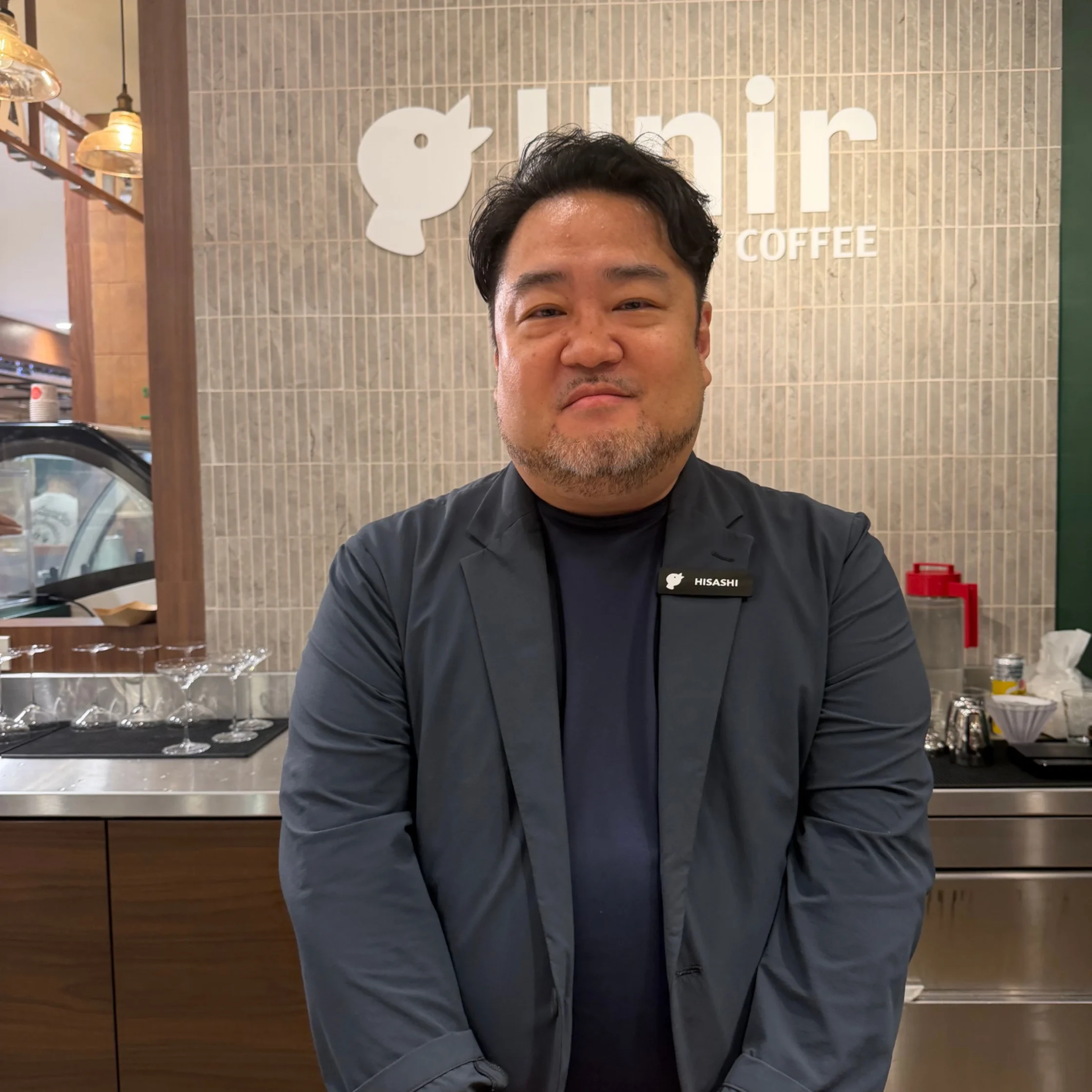 Smiling man in a dark blazer stands in a coffee shop, with a tiled wall and a HISASHI COFFEE logo behind him.