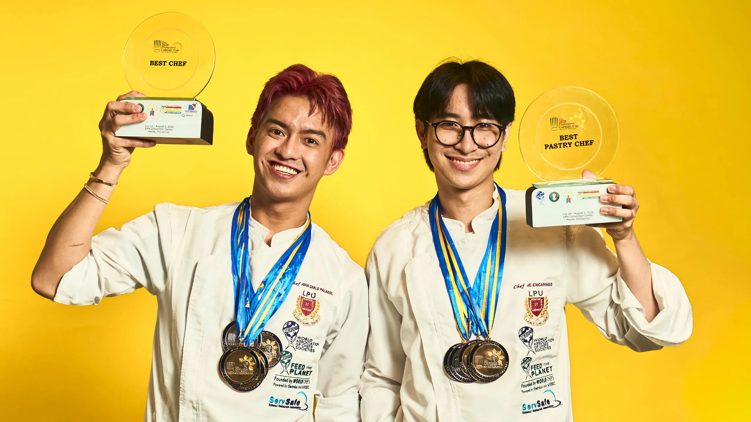 Two chefs in white coats wear blue ribbons and smile, holding large trophy plaques and many medals against a bright yellow background for a celebratory photo. Best Chef of the Year chef John Carlo Palacol and Best Pastry Chef of the Year Jeriel Encarnado of LPU Laguna