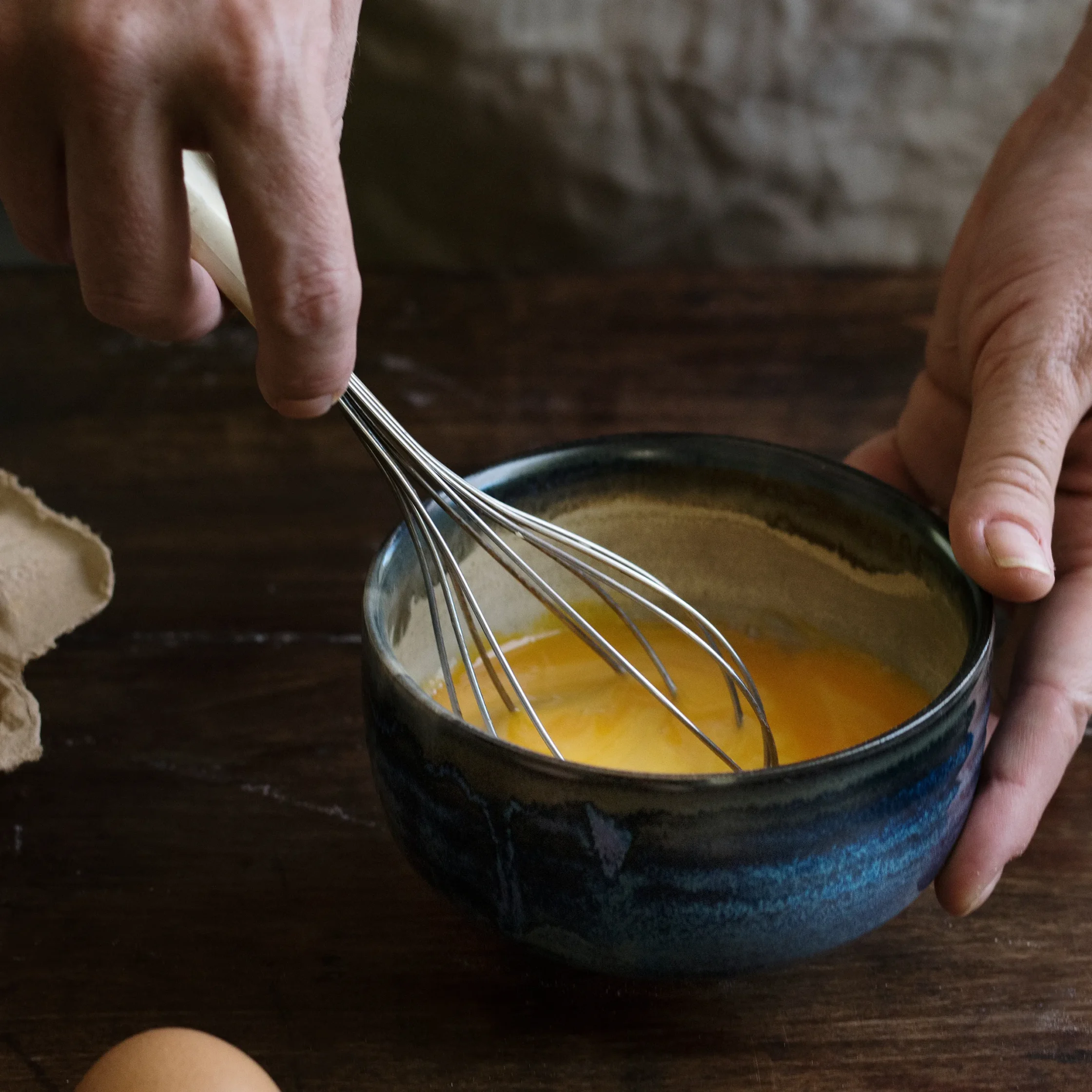 Person's hands whisking yellow batter in a dark ceramic bowl on a wooden table, with a torn bread slice nearby