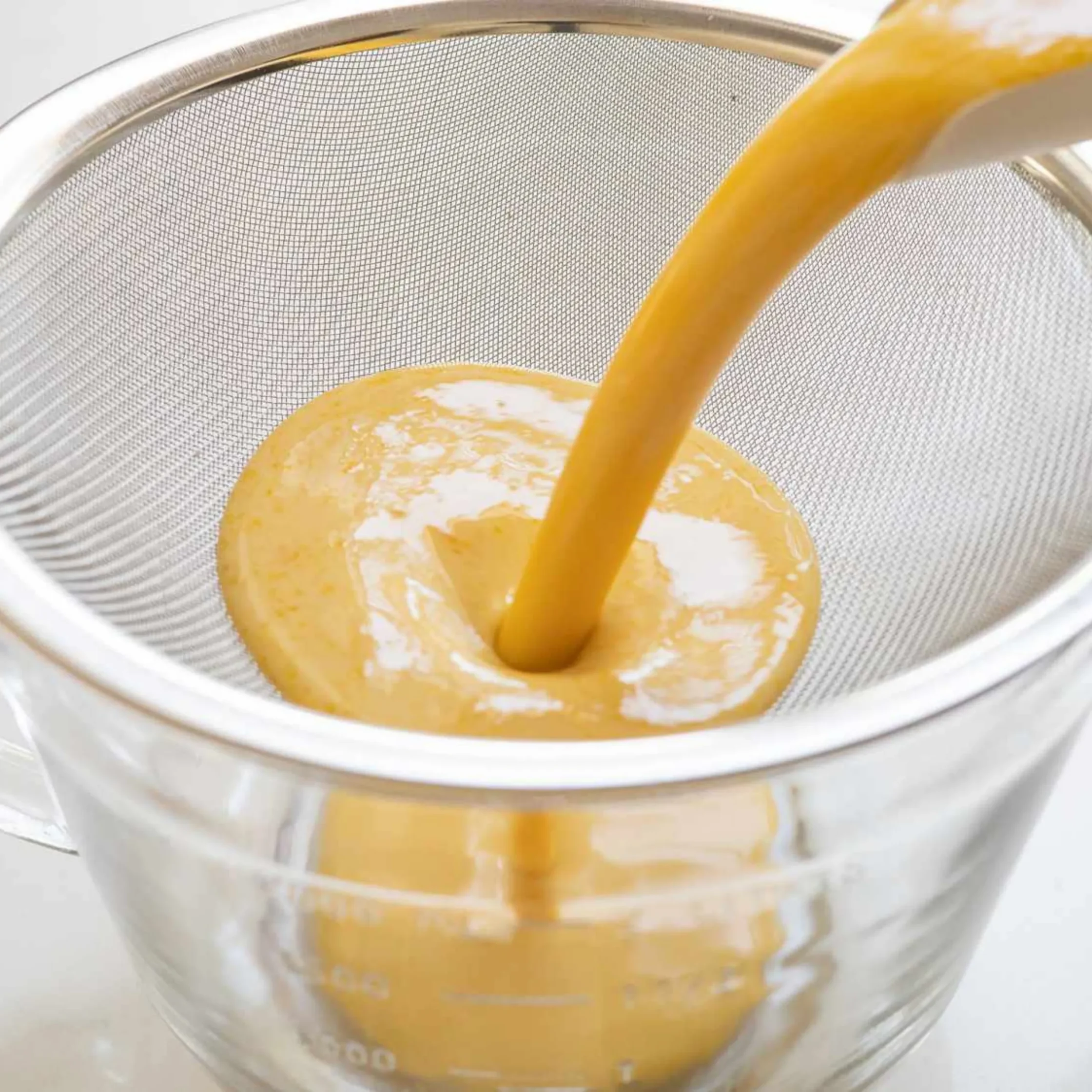 Close-up of orange juice being poured into a glass, with a metal sieve visible in the background.