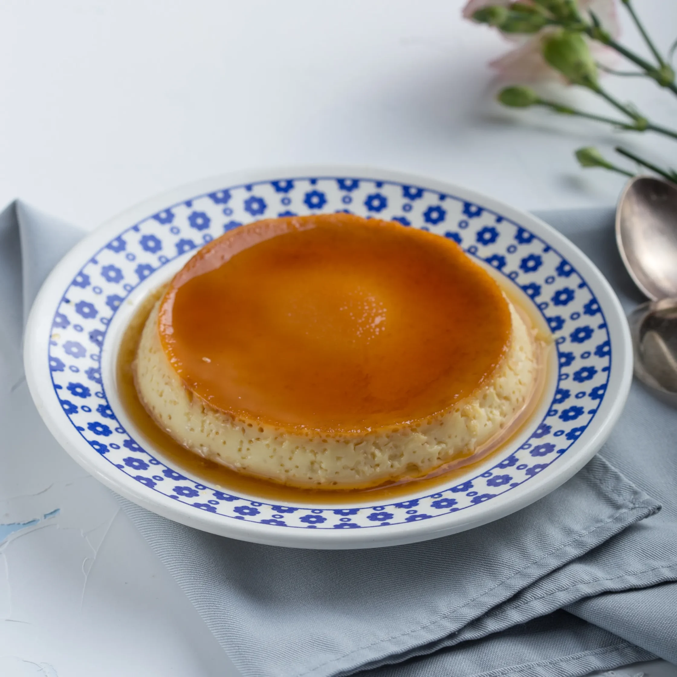 Caramel-topped flan on a blue floral-patterned plate with a spoon nearby on a pale surface.