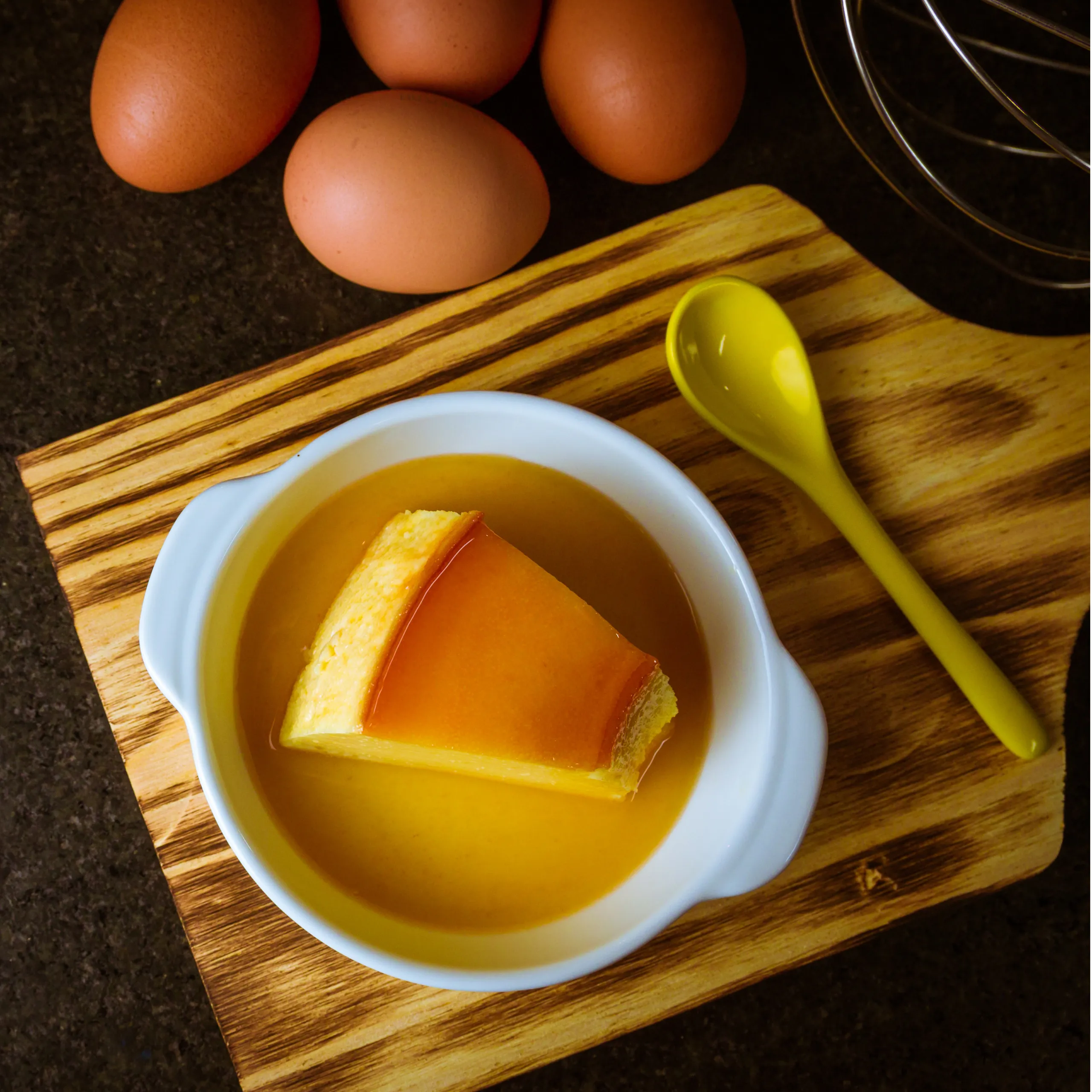 Brown eggs sit on a dark kitchen counter beside a striped wooden cutting board, with a yellow spoon and a bowl containing egg liquid.