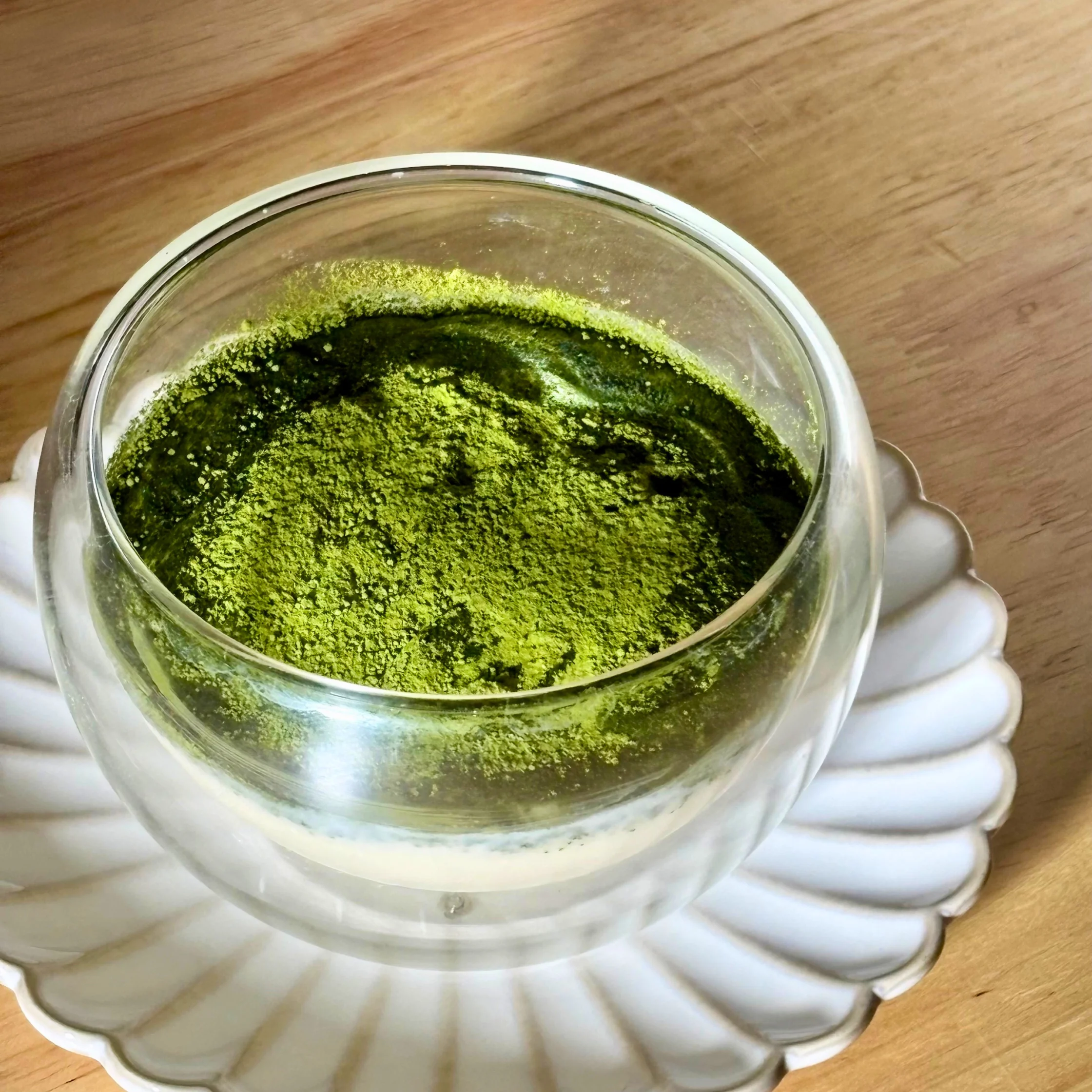 Metal container with bright green powder (likely ground herbs) inside, resting on a wooden surface, gloved hands visible at bottom edge.