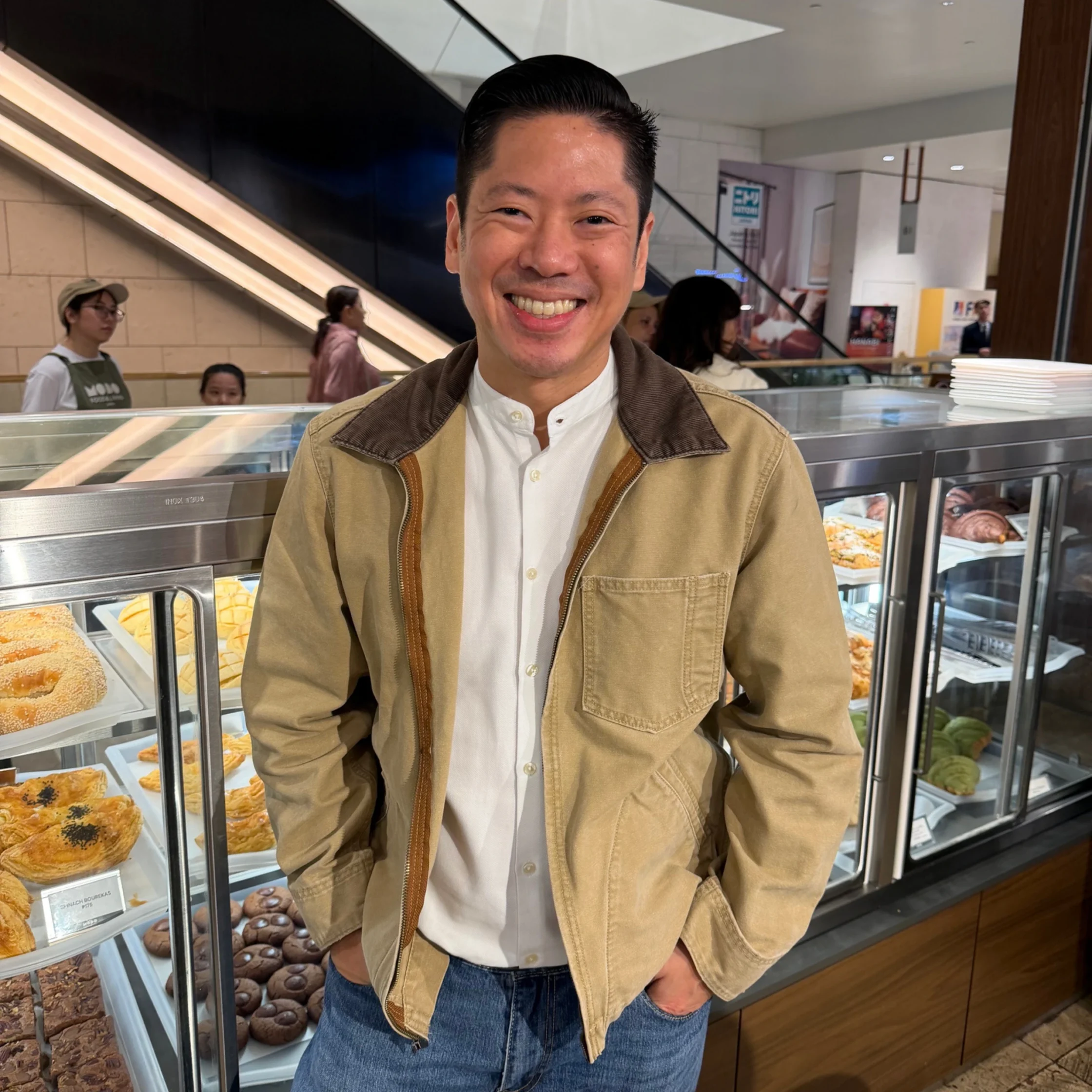 Smiling man in a beige jacket stands at a bakery counter with pastries displayed in a glass case behind him.