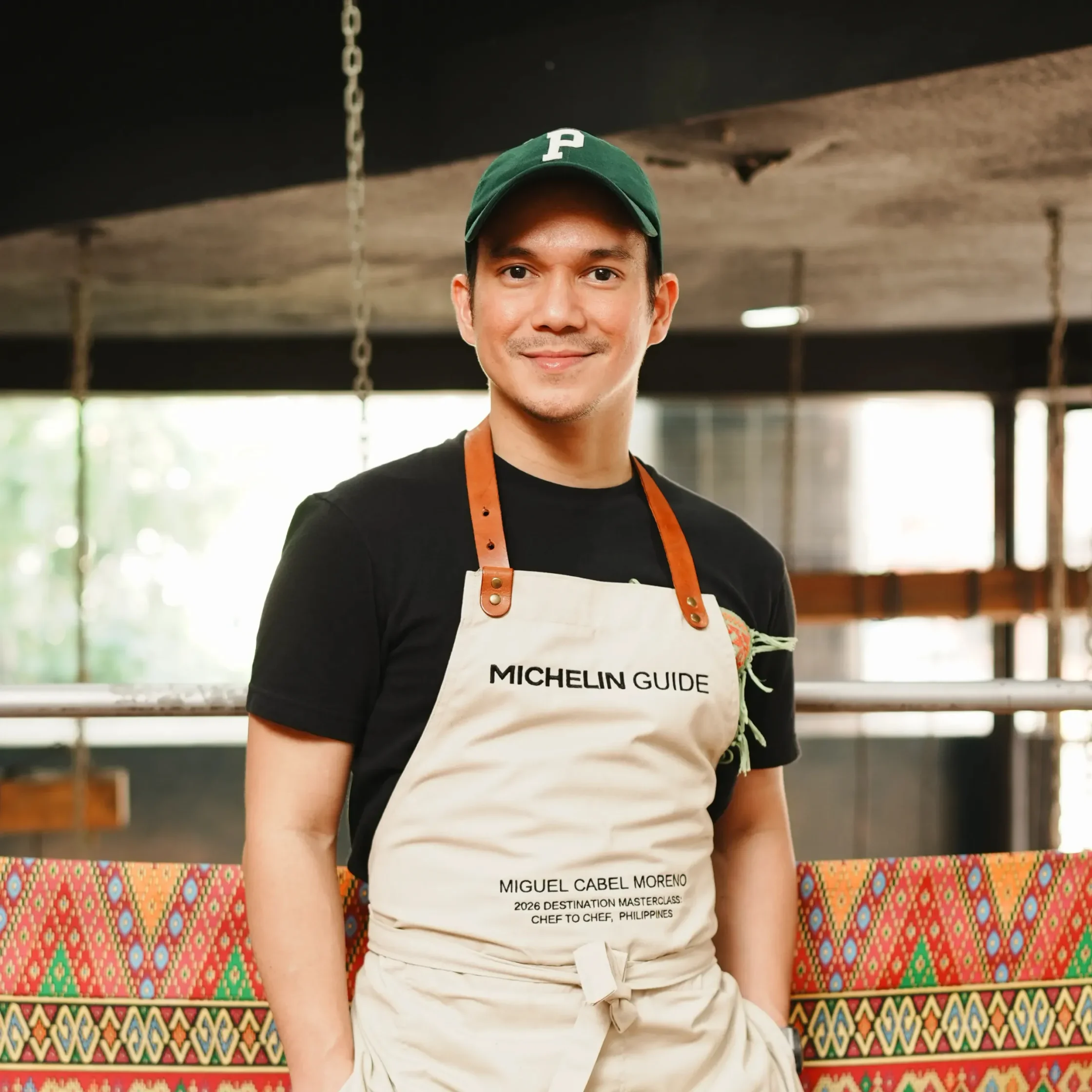 Man wearing a beige Michelin Guide apron and a green cap, standing in front of a colorful patterned bench inside a kitchen or cafe setting.