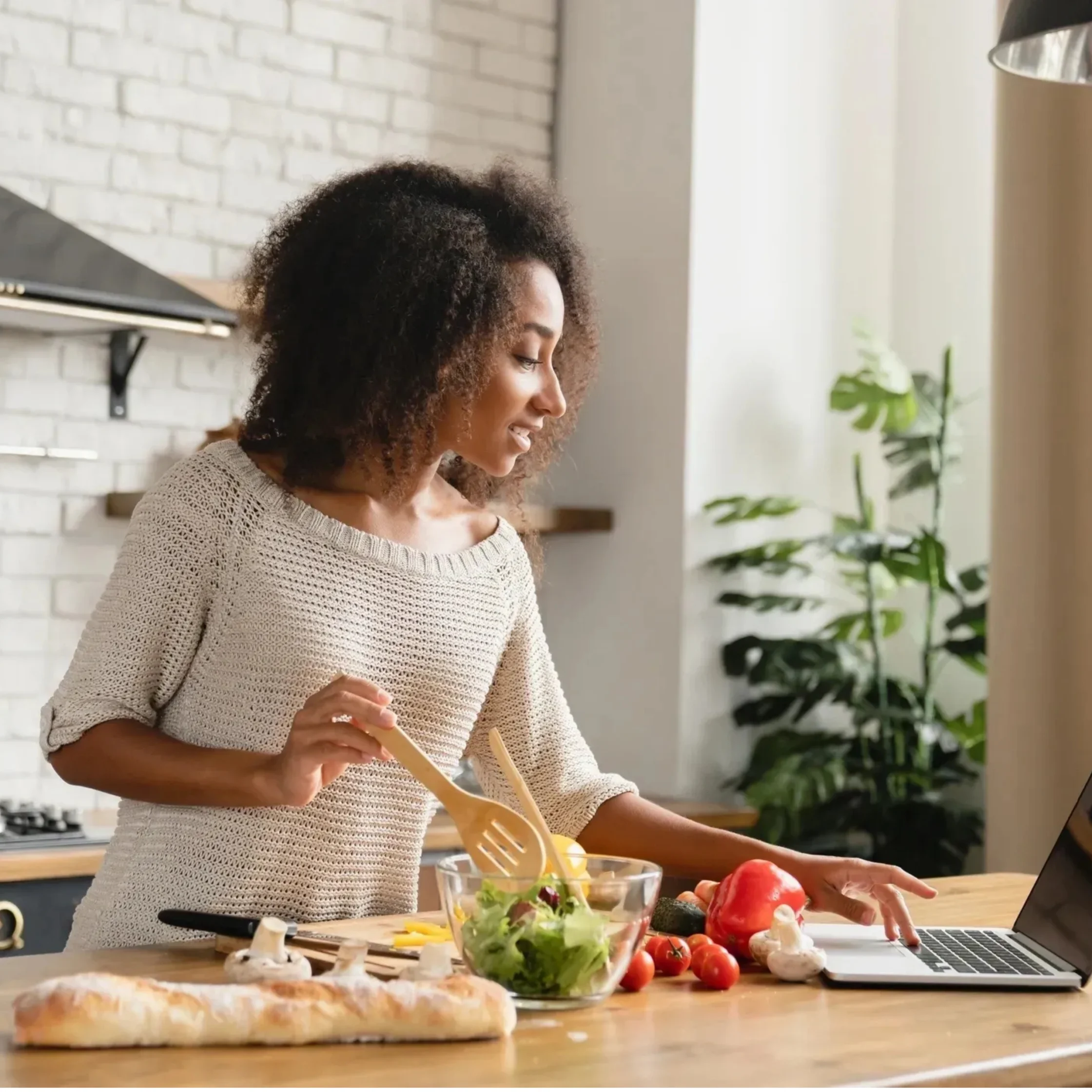 Smiling woman chopping vegetables at a kitchen island with a laptop nearby and a salad bowl in front of her.