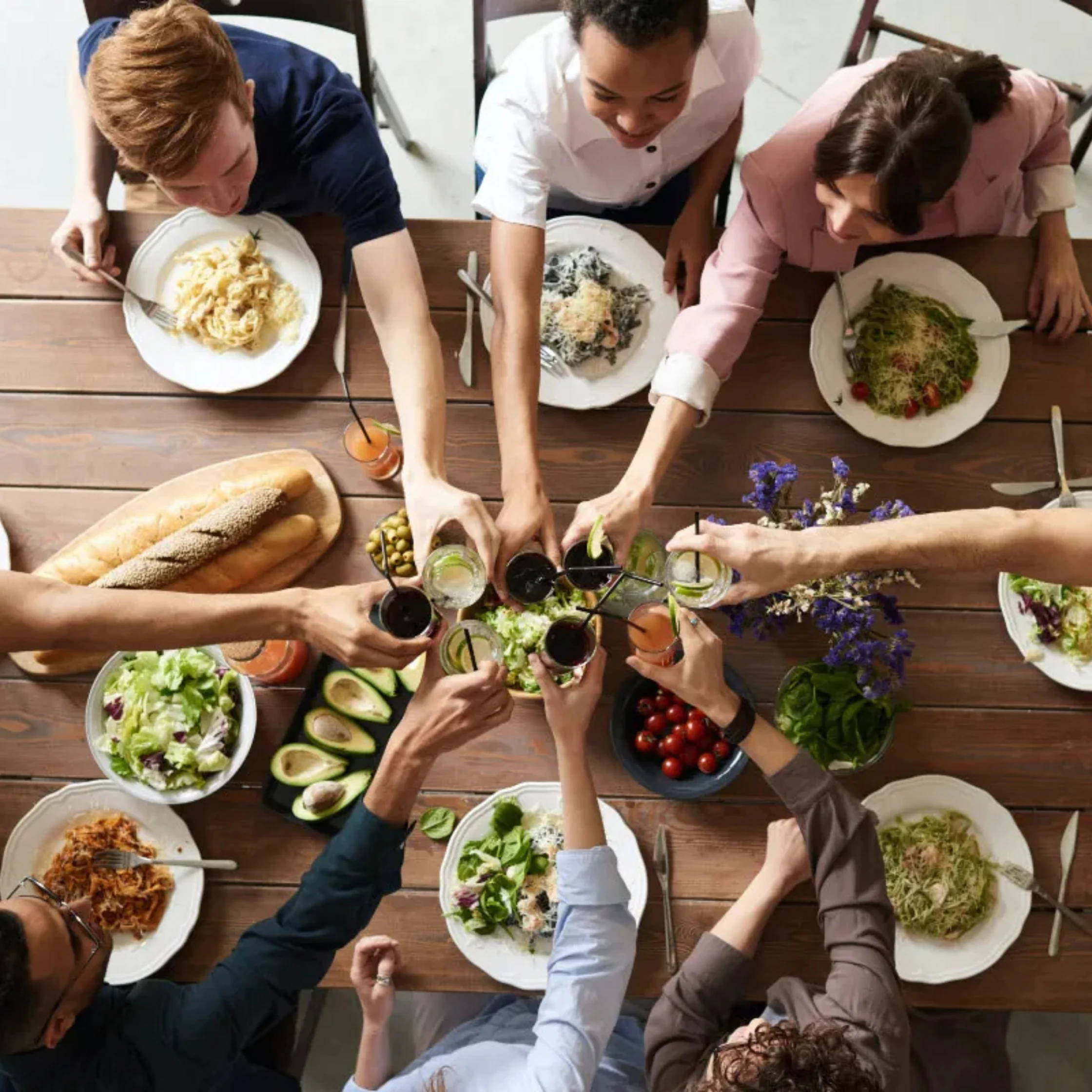 Top-down view of diverse friends around a wooden table clinking glasses as they share a meal of salad, bread, pasta, and toppings.