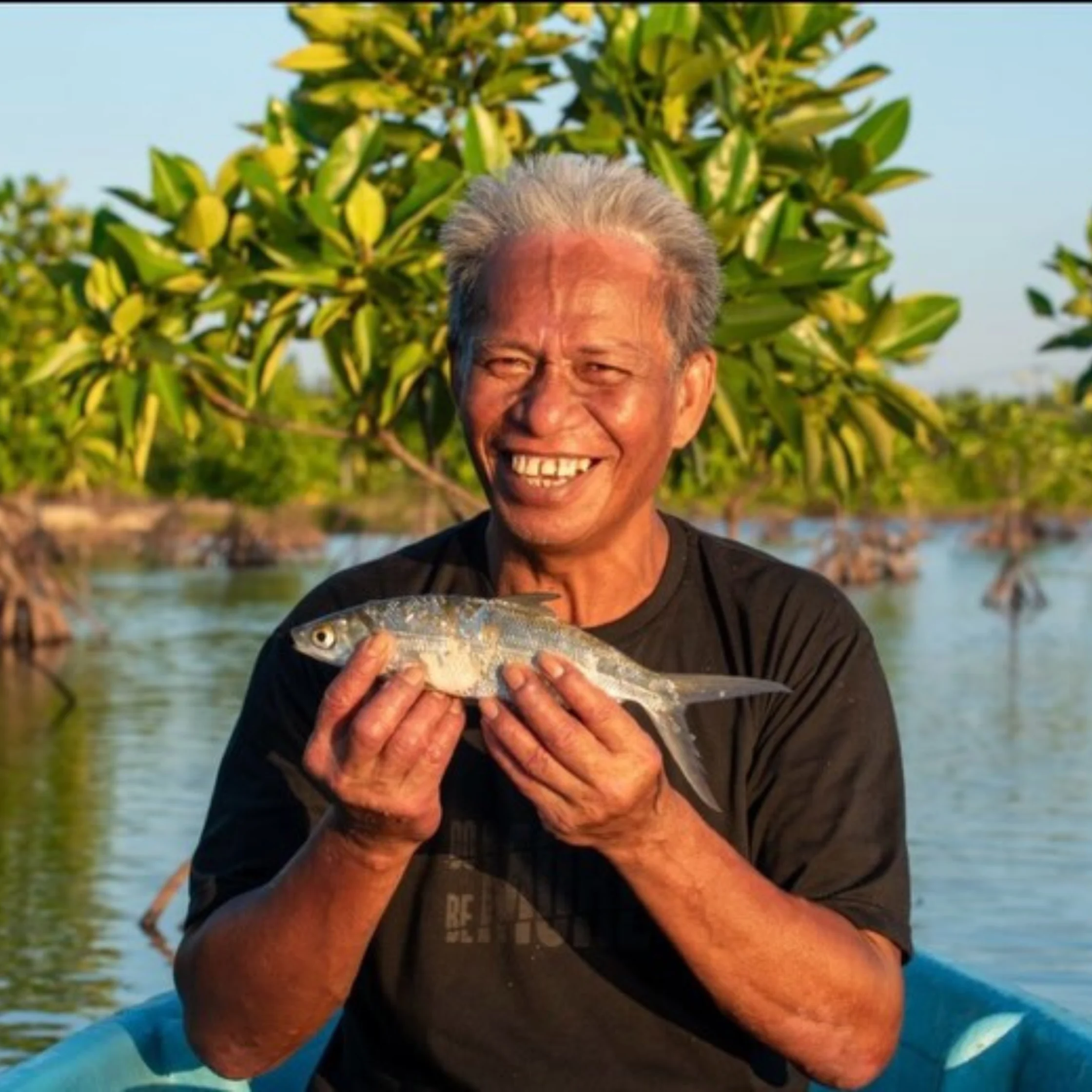Smiling older man holding a silver fish on a boat by calm water with green trees in the background.
