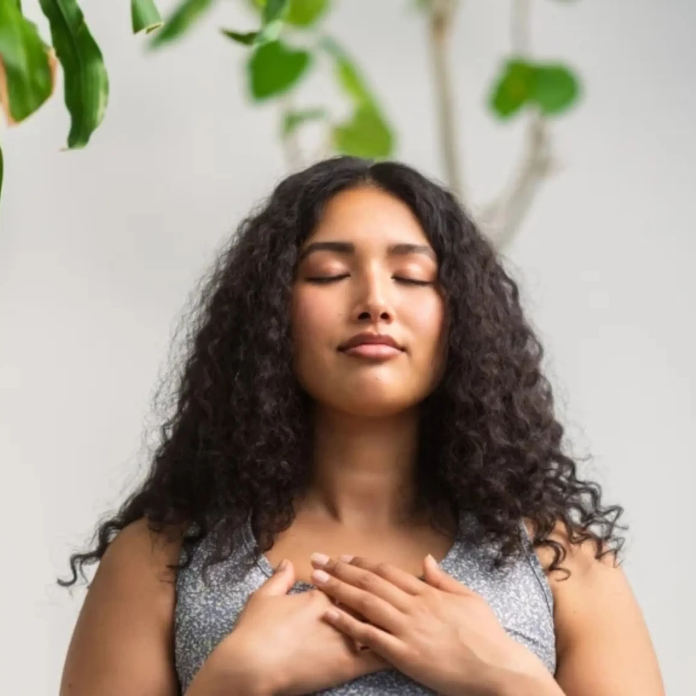 Young woman with curly hair, eyes closed, hands crossed over her chest in a calm indoor pose.