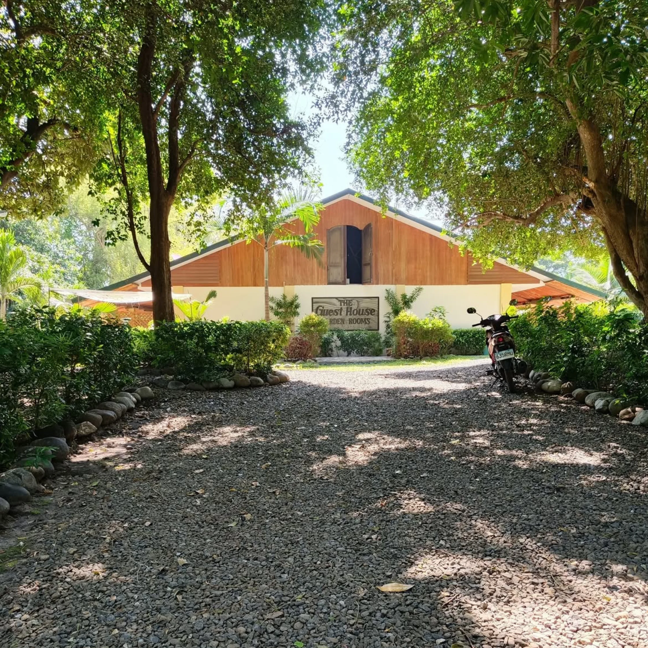 Gravel driveway leading to a wooden house framed by leafy trees; a scooter parked to the right and a sign reading 'The Guest House' visible near the entrance.