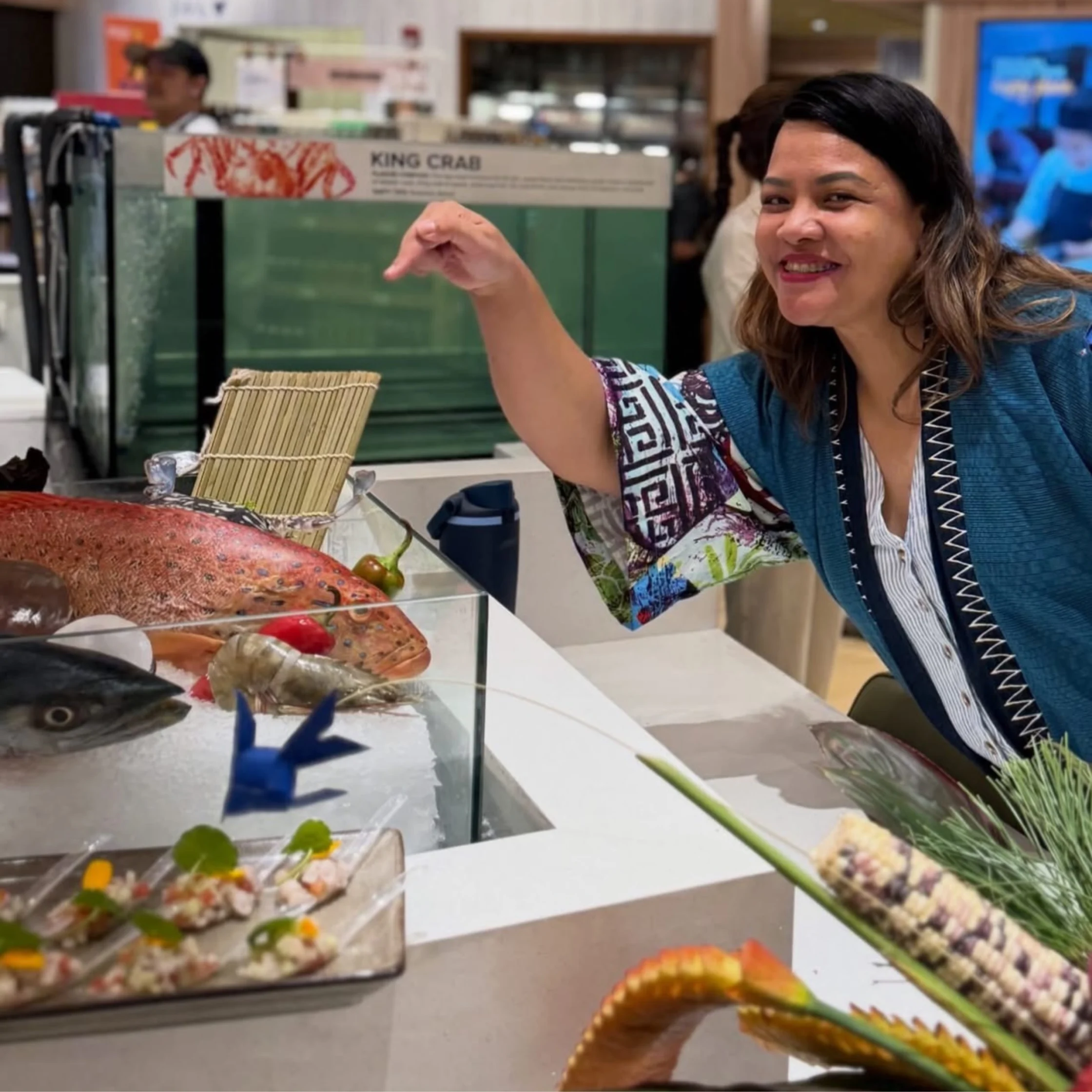 Smiling woman at a seafood counter pointing toward a display of crab legs and other seafood behind glass, with a 'King Crab' sign in the background.