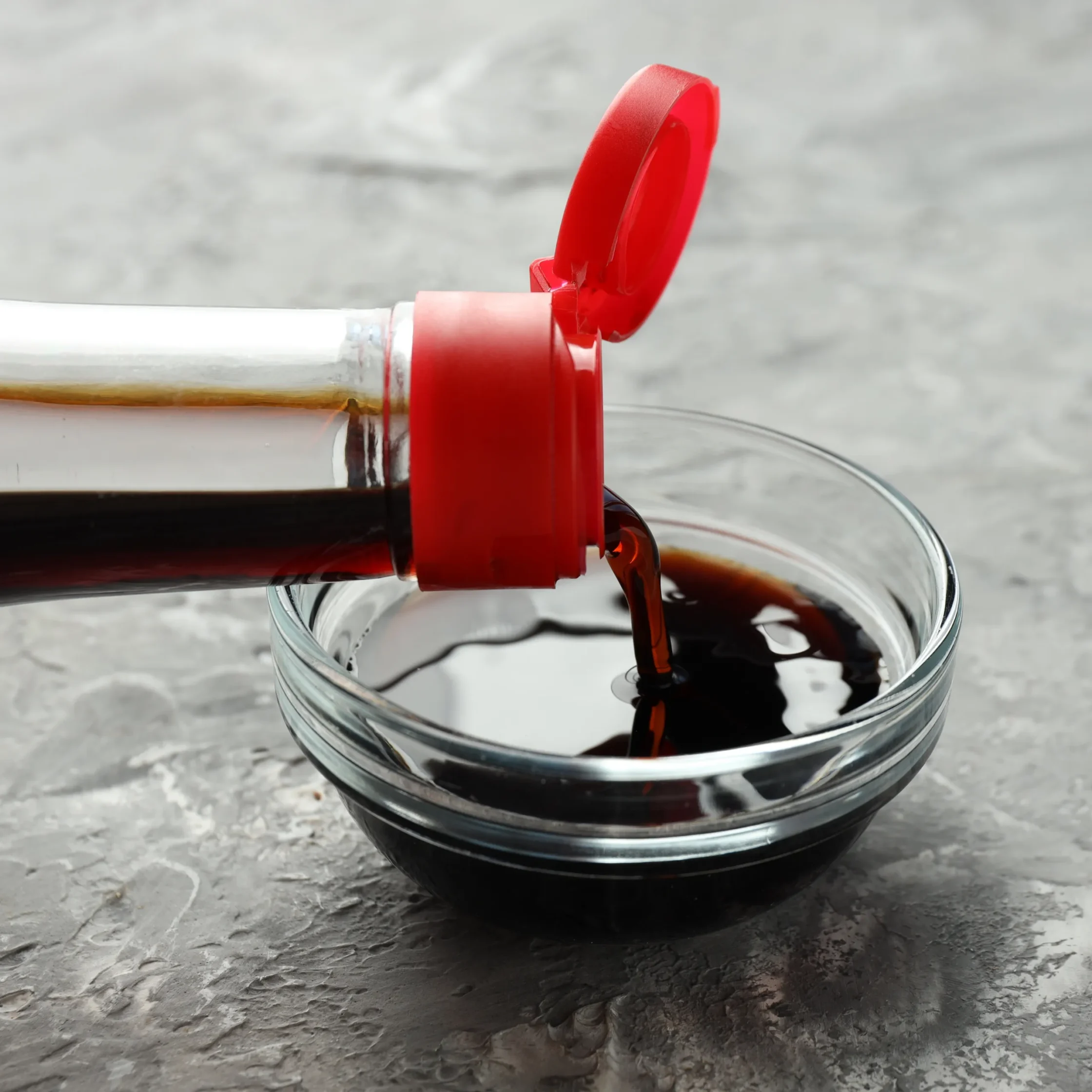 Red lipstick case tipped over a clear glass as red liquid pours into it on a textured gray surface.