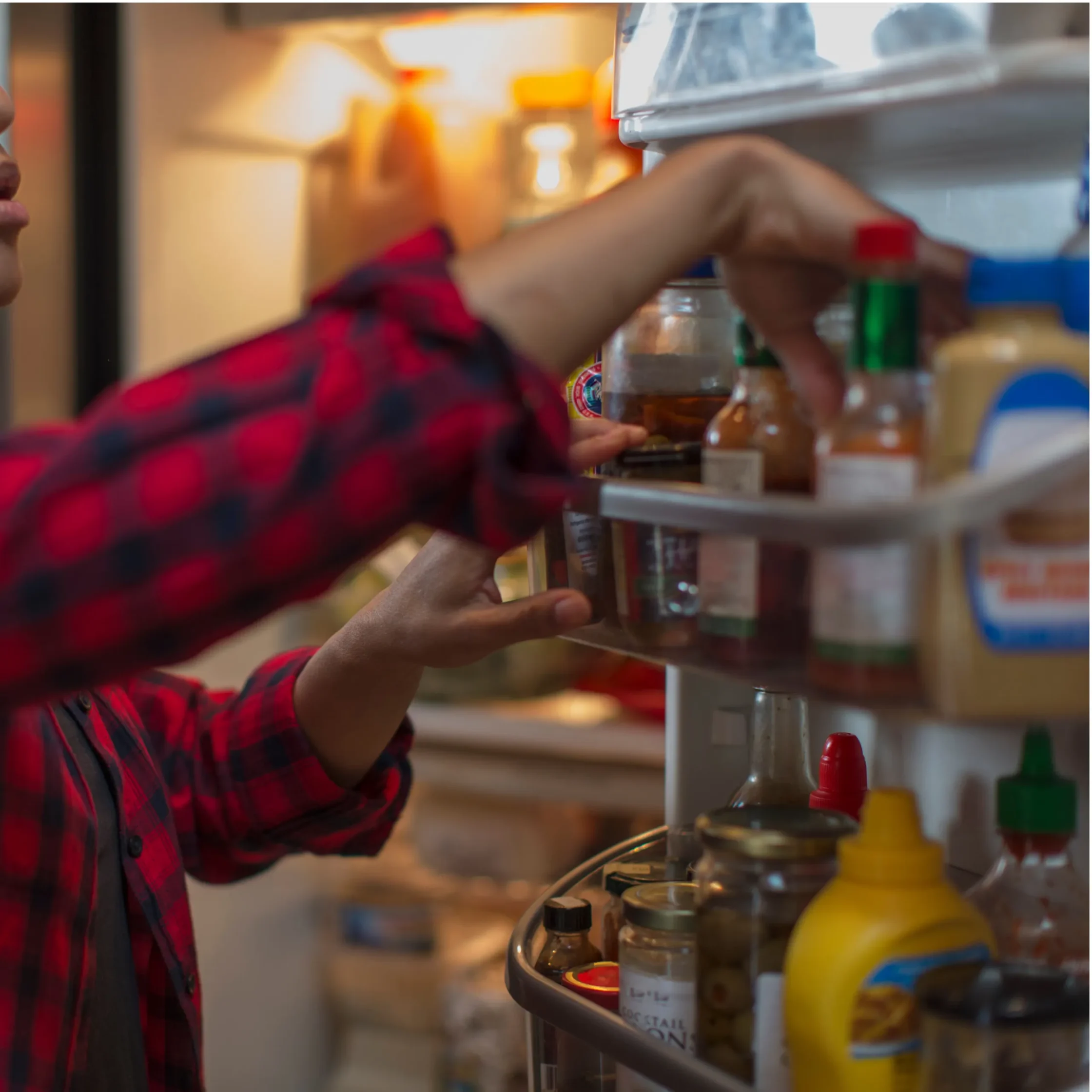 Person wearing a red plaid shirt reaching into a refrigerated display case, grabbing a bottle from shelves of sauces and condiments in a grocery store.
